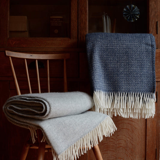 A styled photograph showing a grey and blue wool blanket, styled on top of a chair and infront of a wooden cabinet.