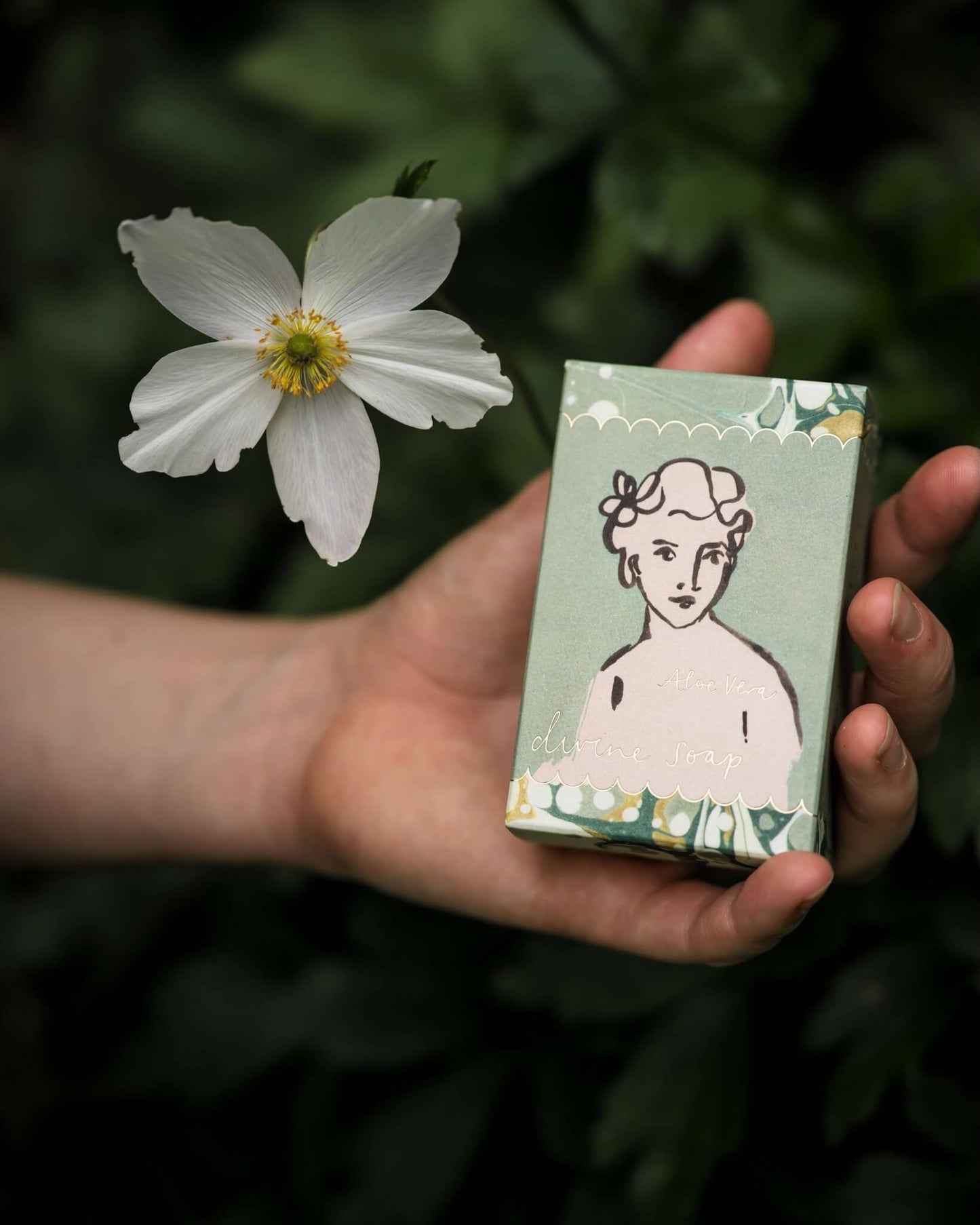 Hand holding a green box of soap with a printed design of a woman's face, next to a white flower against a green background.