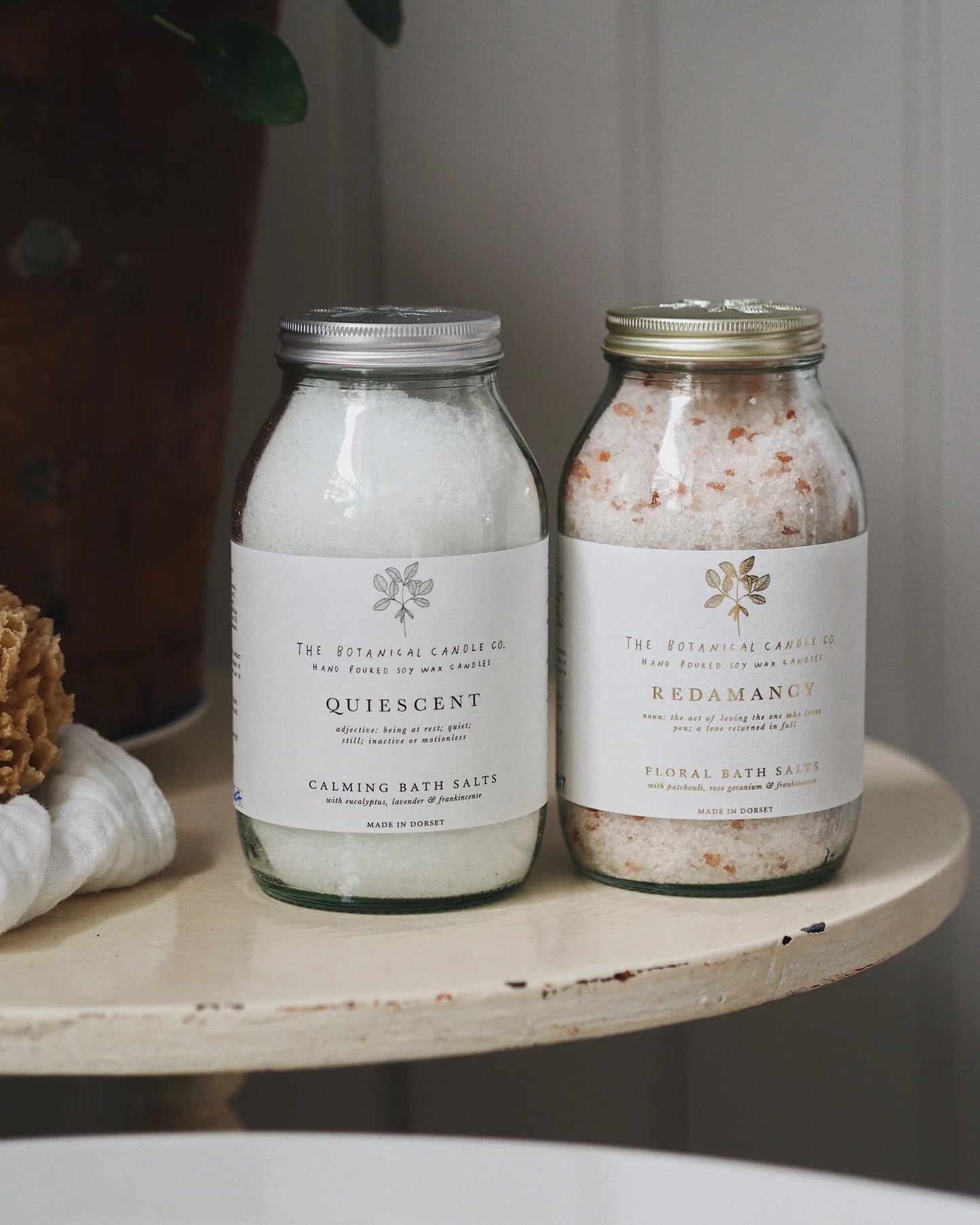 Two jars of bath salts labeled 'Quiescent' and 'Redamancy' on a wooden bathroom surface.