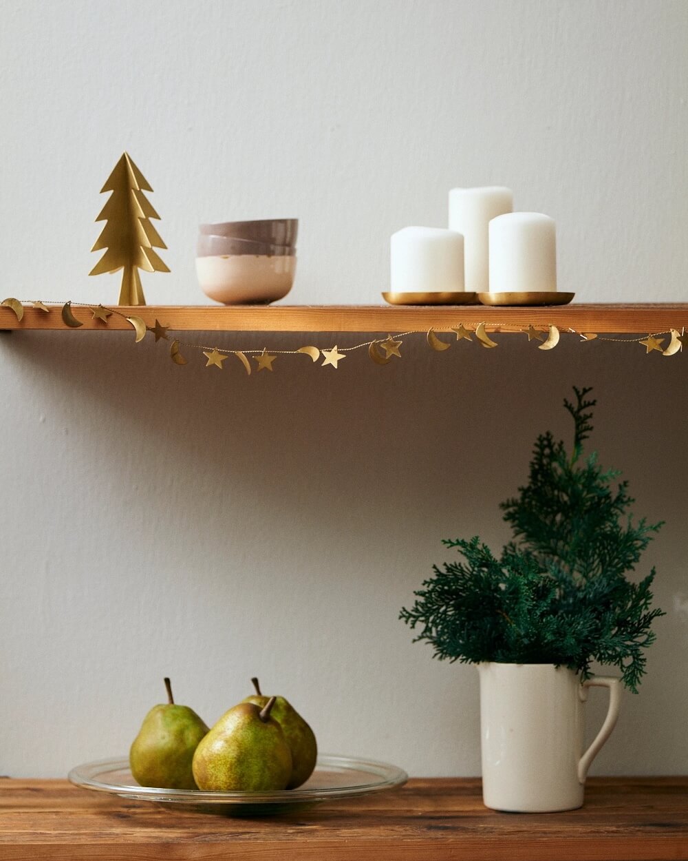 Decorative shelf with brass Christmas garland, tree, bowls, candles, and a plant on a wooden surface.