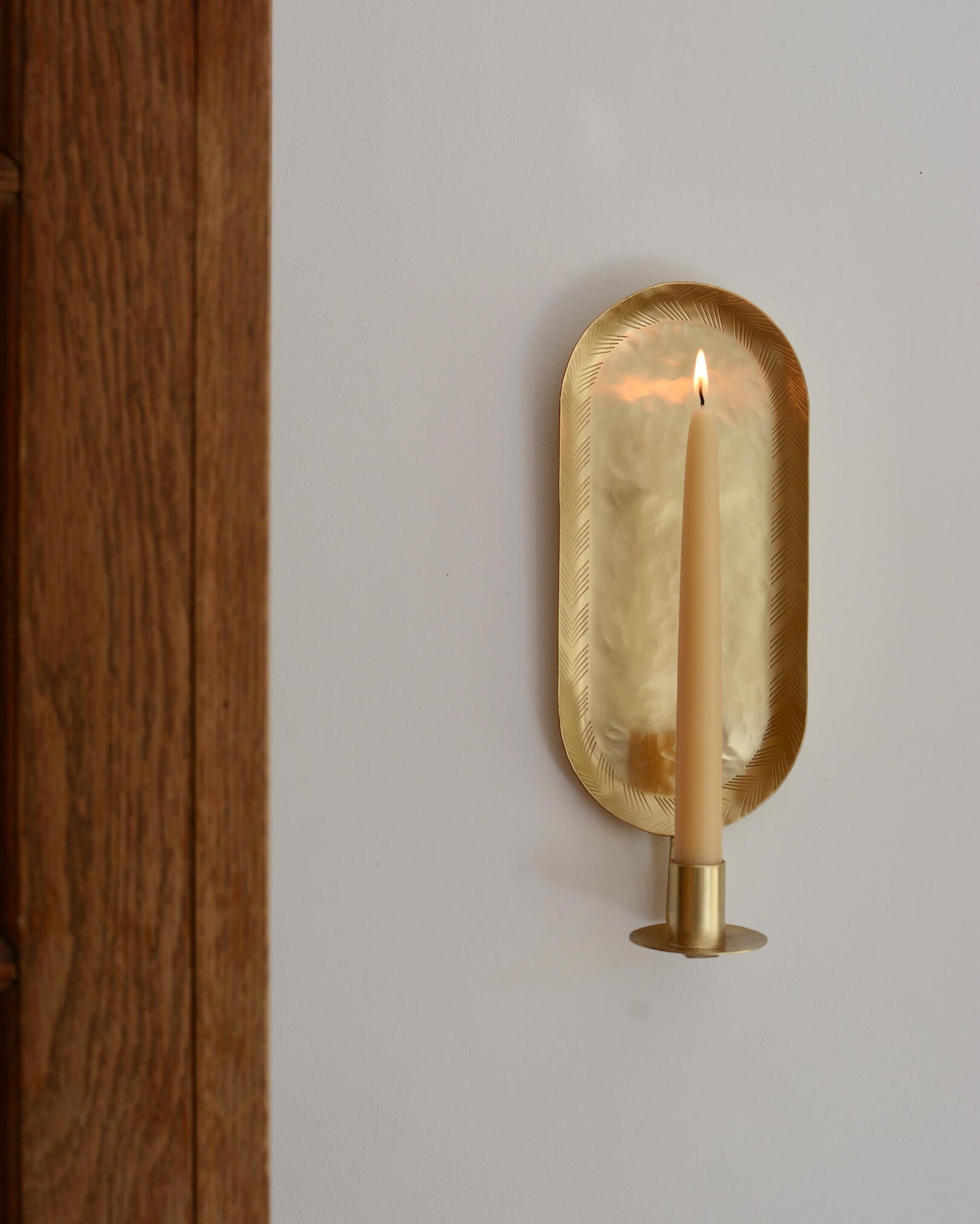 A brass candle wall sconce in an oval shape, with a lit beeswax taper candle.