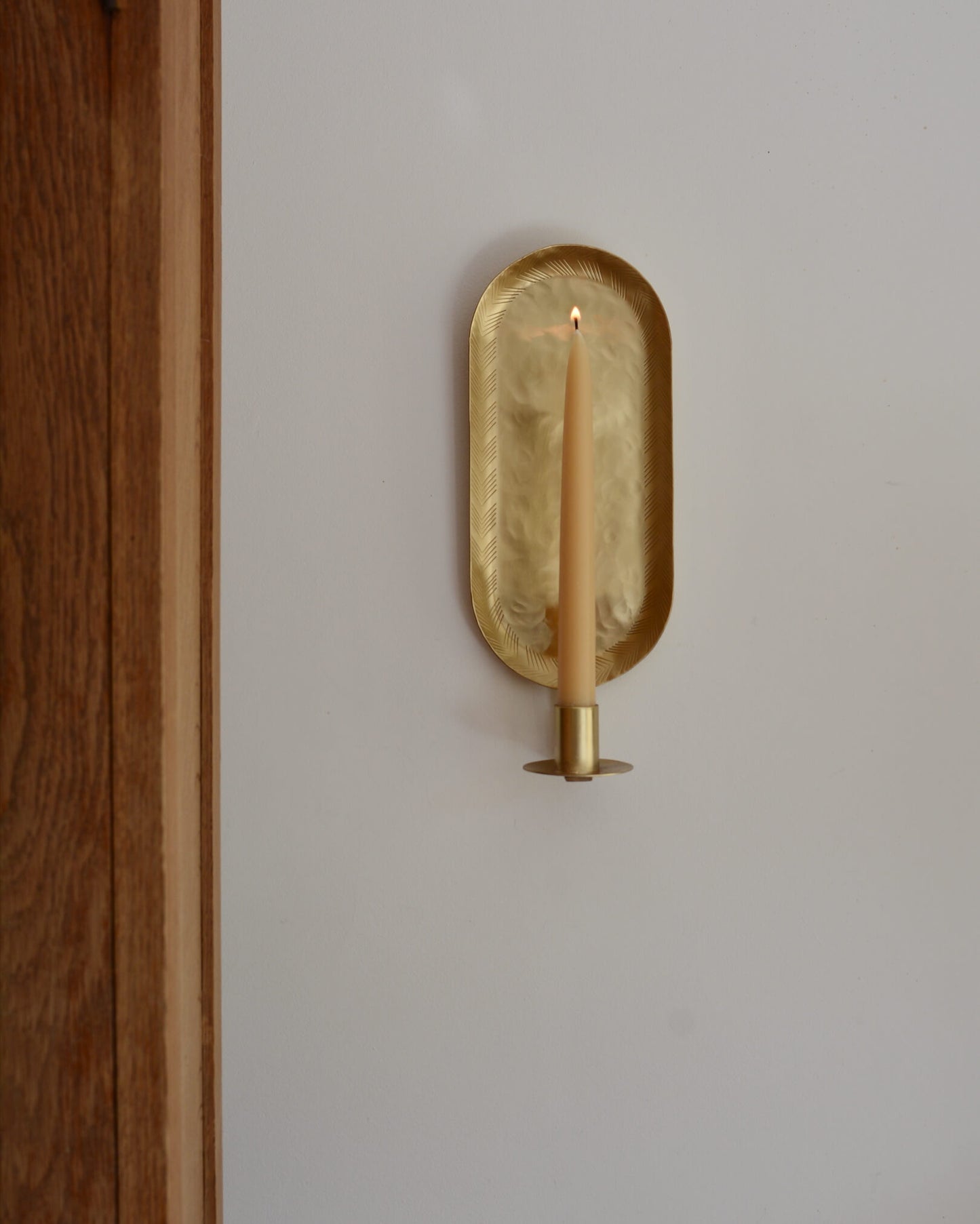 A brass candle wall sconce in an oval shape, with a lit beeswax taper candle.