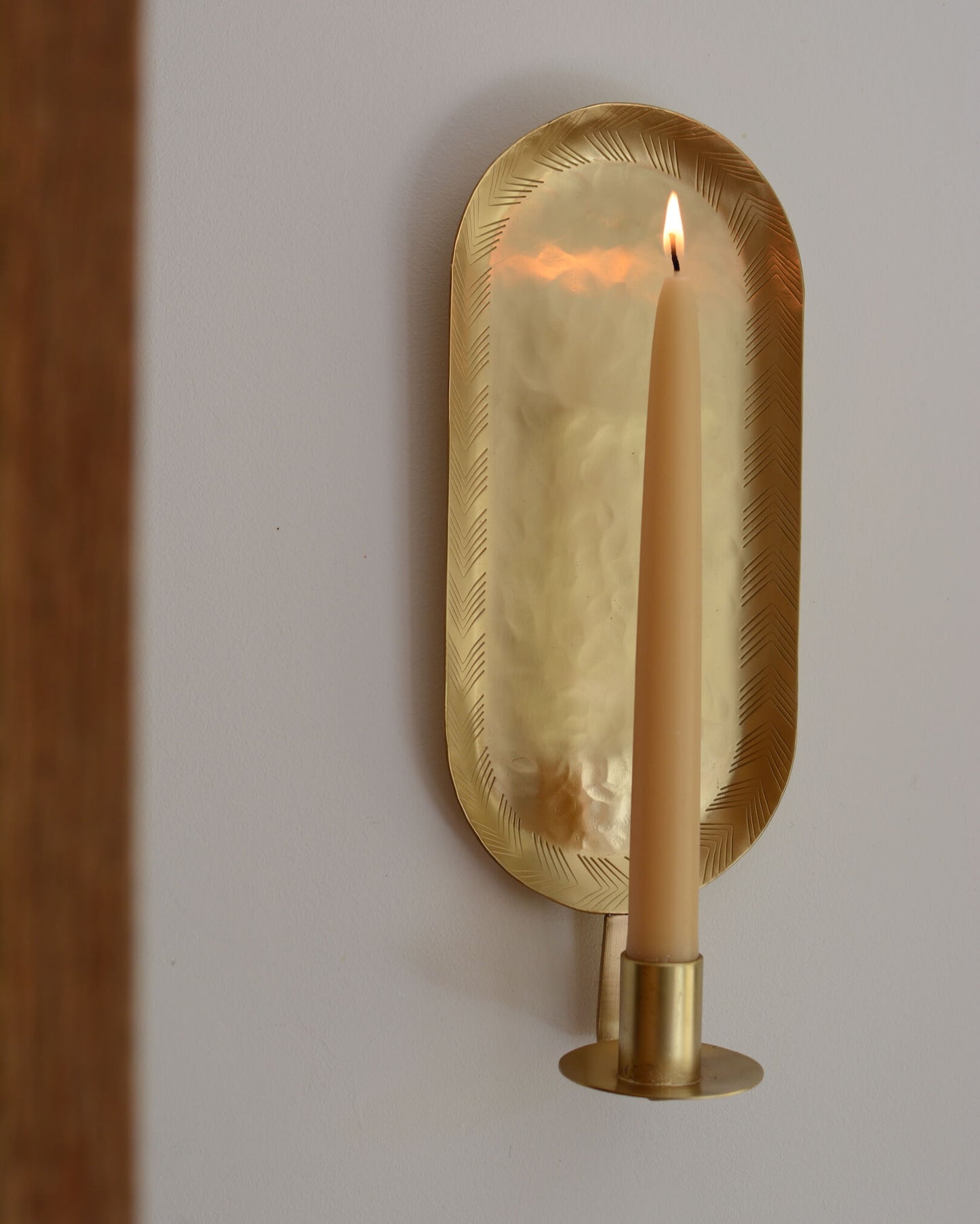 A brass candle wall sconce in an oval shape, with a lit beeswax taper candle.