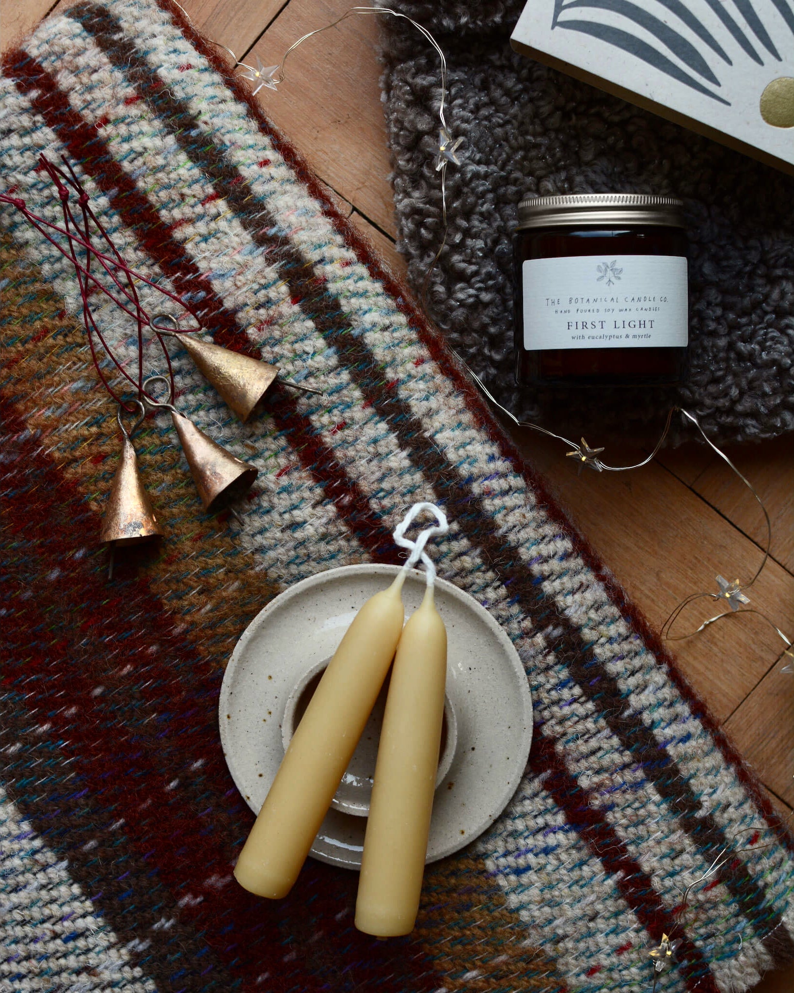 A pair of pure beeswax short taper candles, next to a set of Christmas bells and a woollen blanket.
