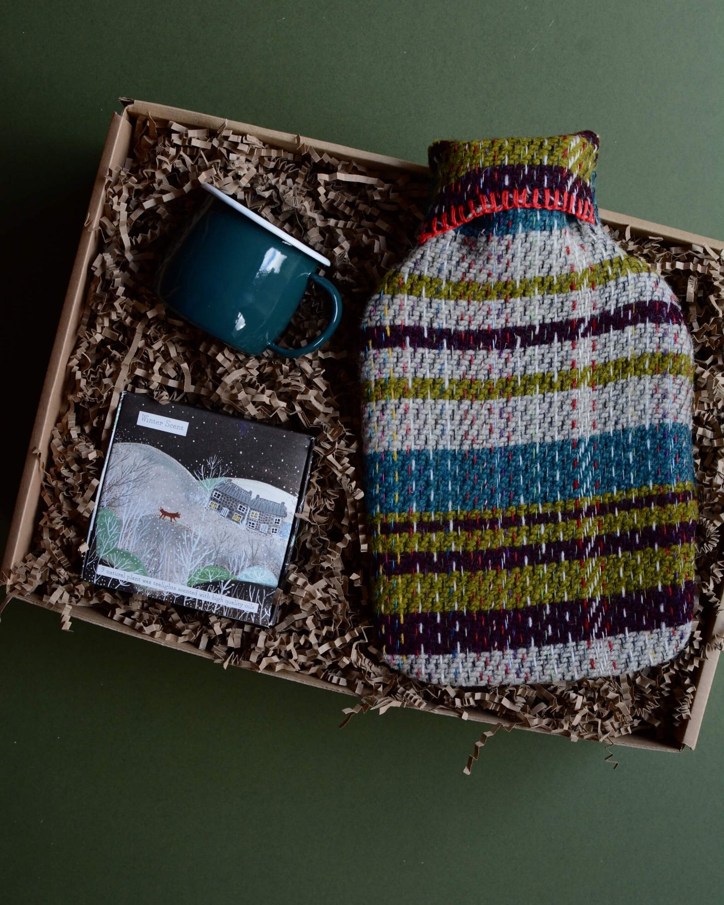 A wool hot water bottle, green enamel mug, and box of tealights in a gift box, shown on a green background.