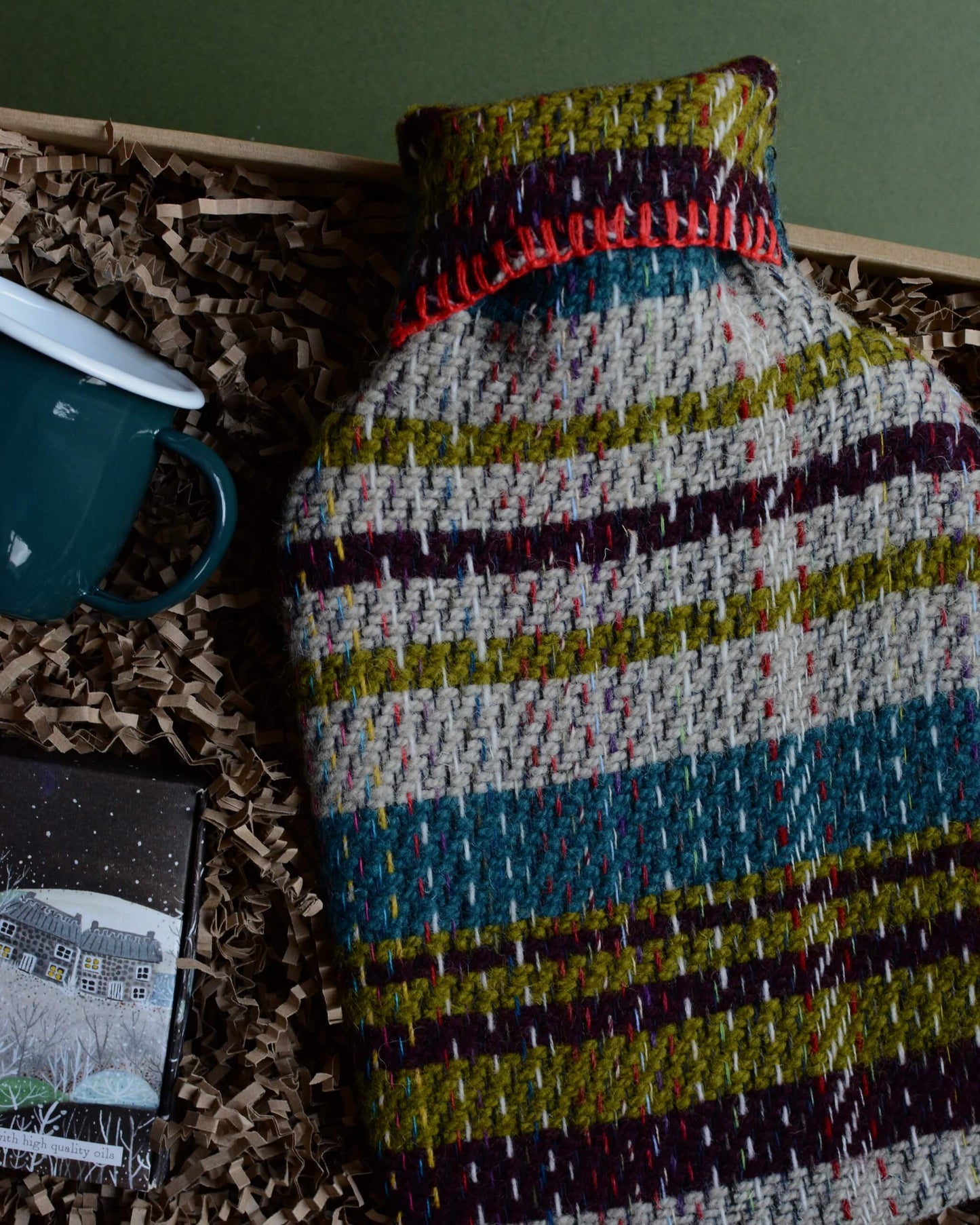 A wool hot water bottle, green enamel mug, and box of tealights in a gift box, shown on a green background.