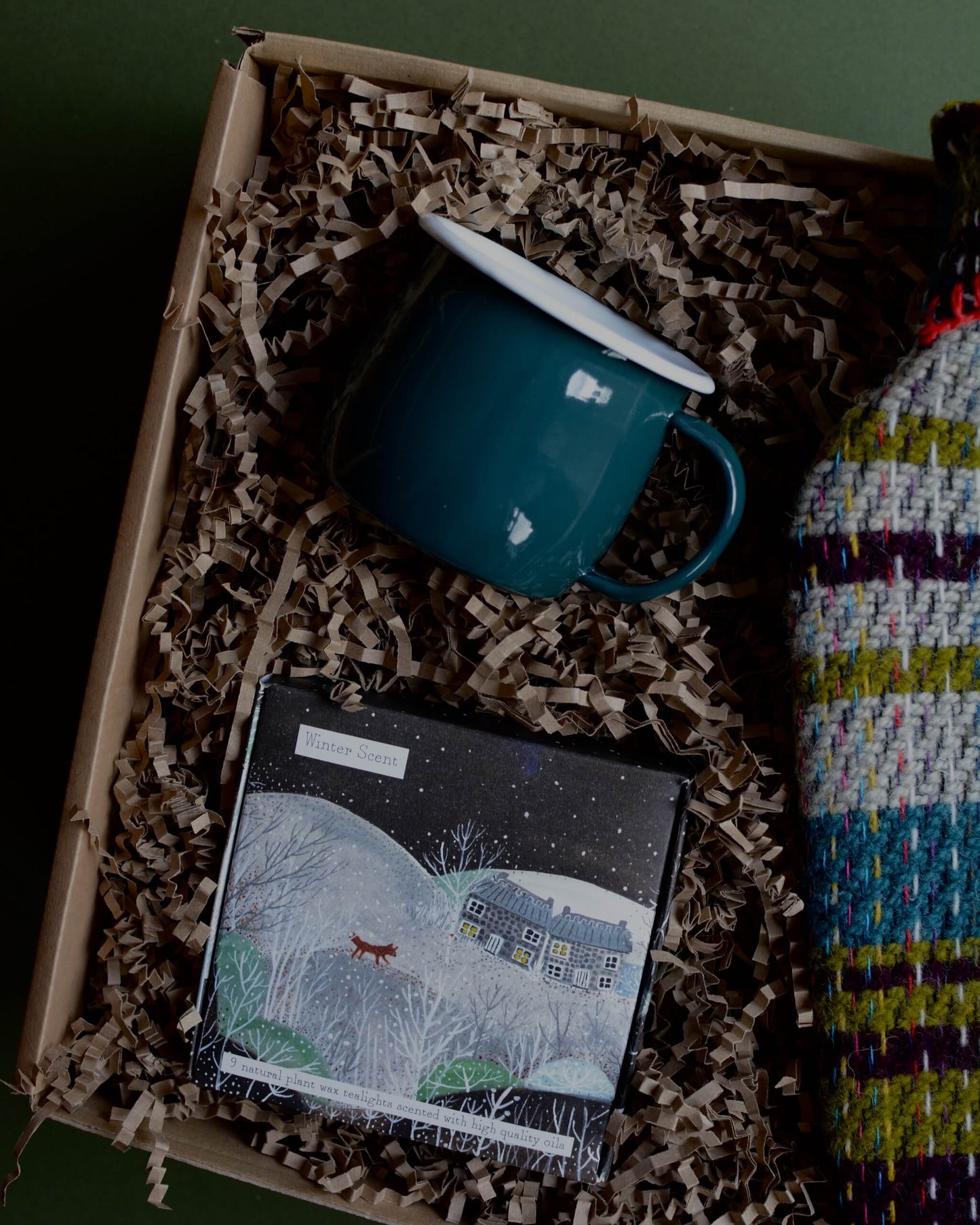 A wool hot water bottle, green enamel mug, and box of tealights in a gift box, shown on a green background.