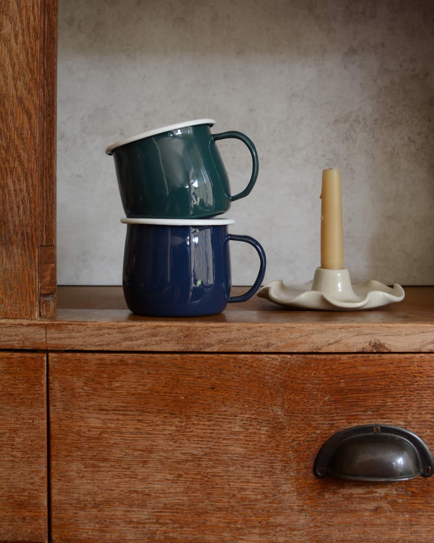 A pair of green and navy blue enamel mugs, alongside a beeswax candle in a ceramic candle holder.
