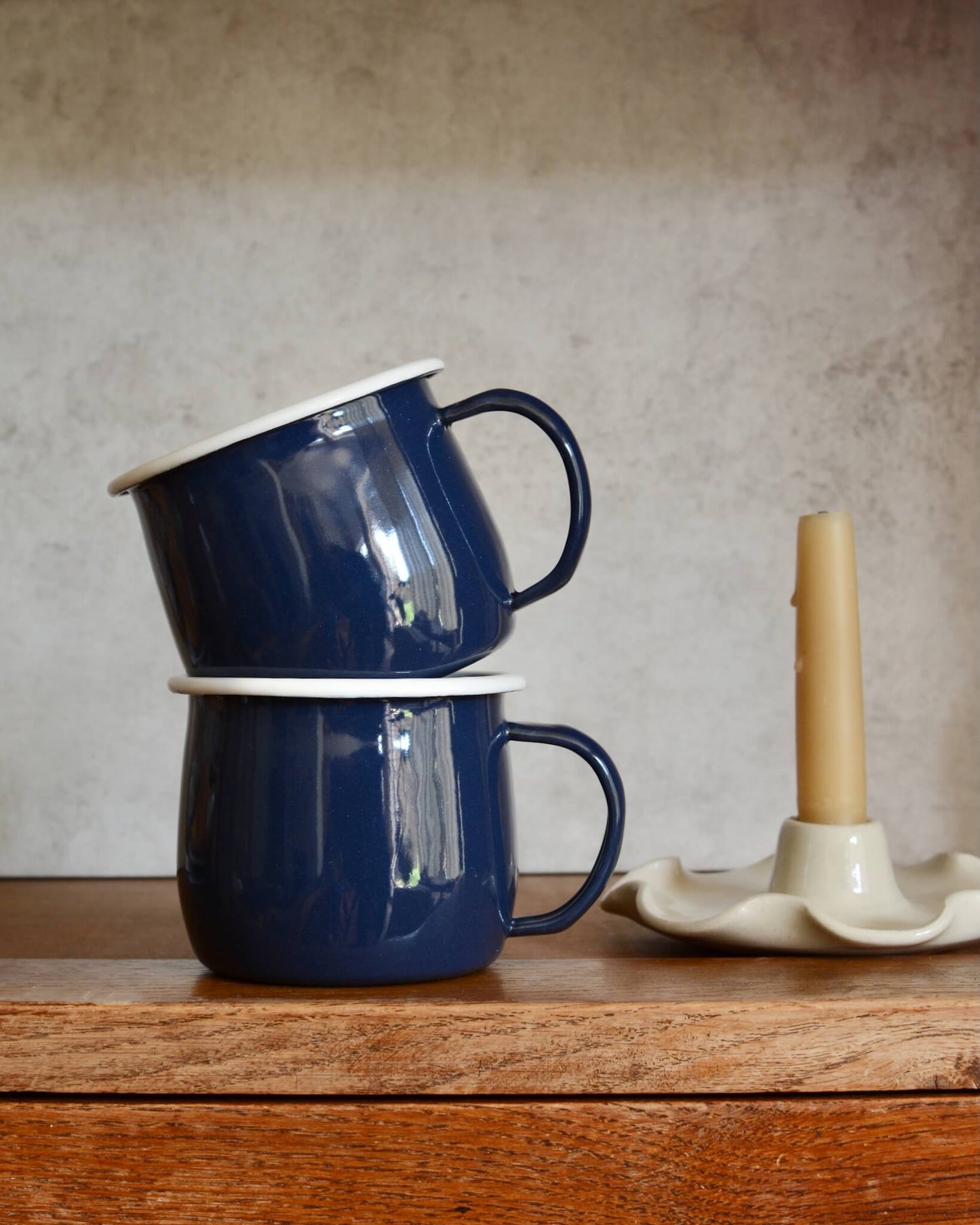 A pair of navy blue enamel mugs, alongside a beeswax candle in a ceramic candle holder.