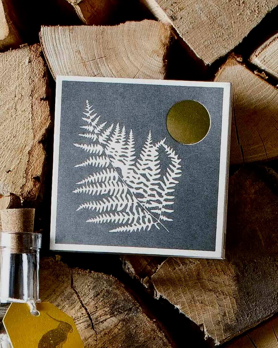 Fern print design matchbox, placed on stacked wooden logs.