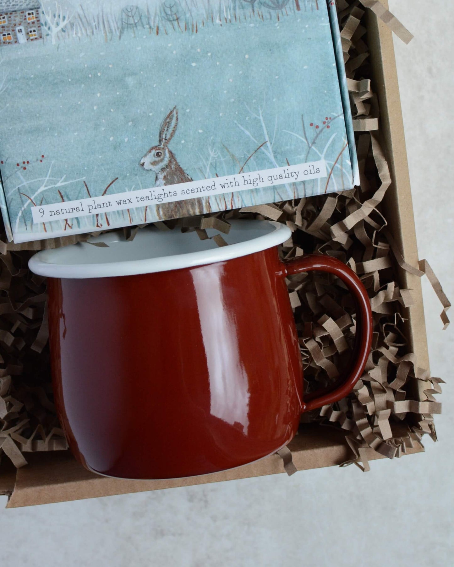 Red mug and illustrated box of tealight candles in a gift box with crinkled paper.