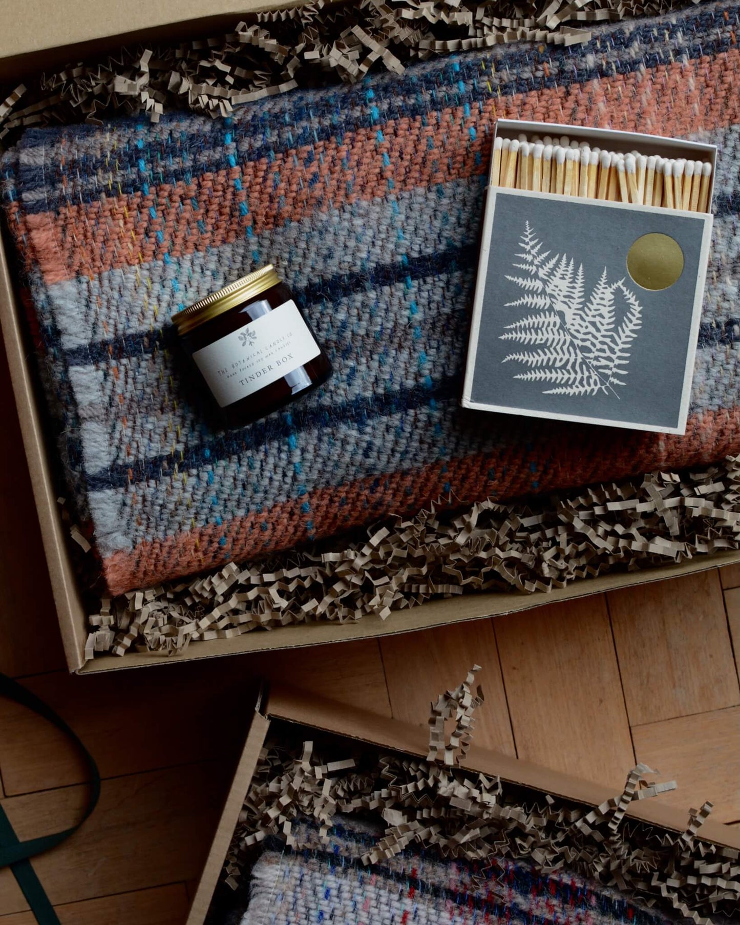 An example of a gift box, containing a checked woollen blanket, a candle and a box of matches.