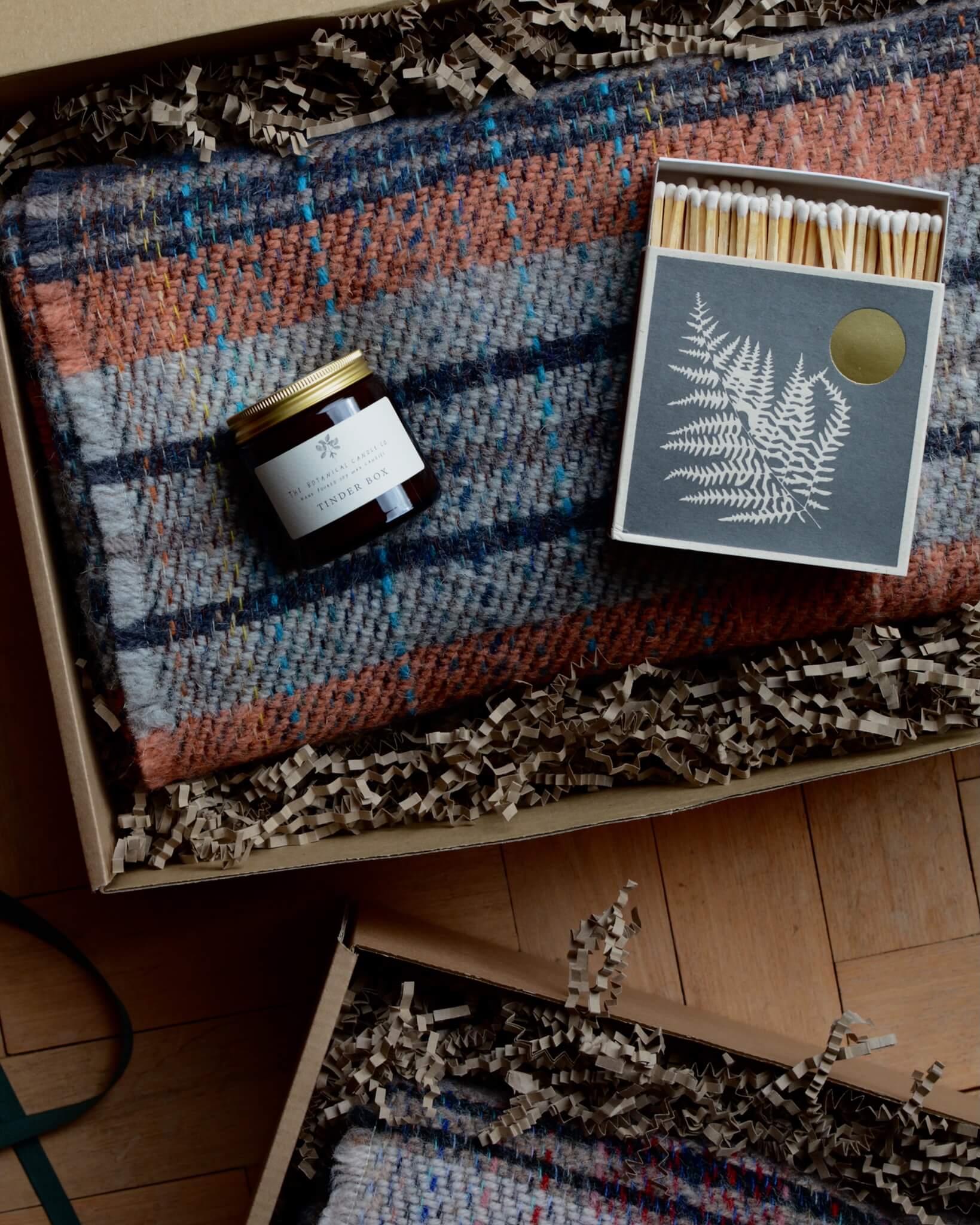 An example of a gift box, containing a checked woollen blanket, a candle and a box of matches.
