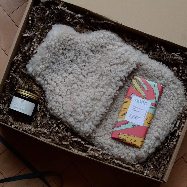 A gift box containing a sheepskin hot water bottle, jar candle and bar of chocolate.
