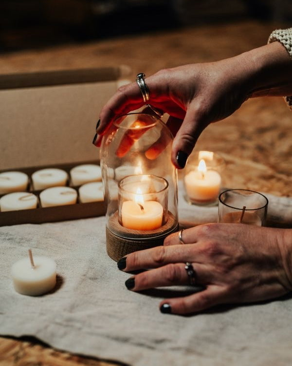 Person holding a glass lantern with a lit candle, surrounded by other candles on a wooden surface.