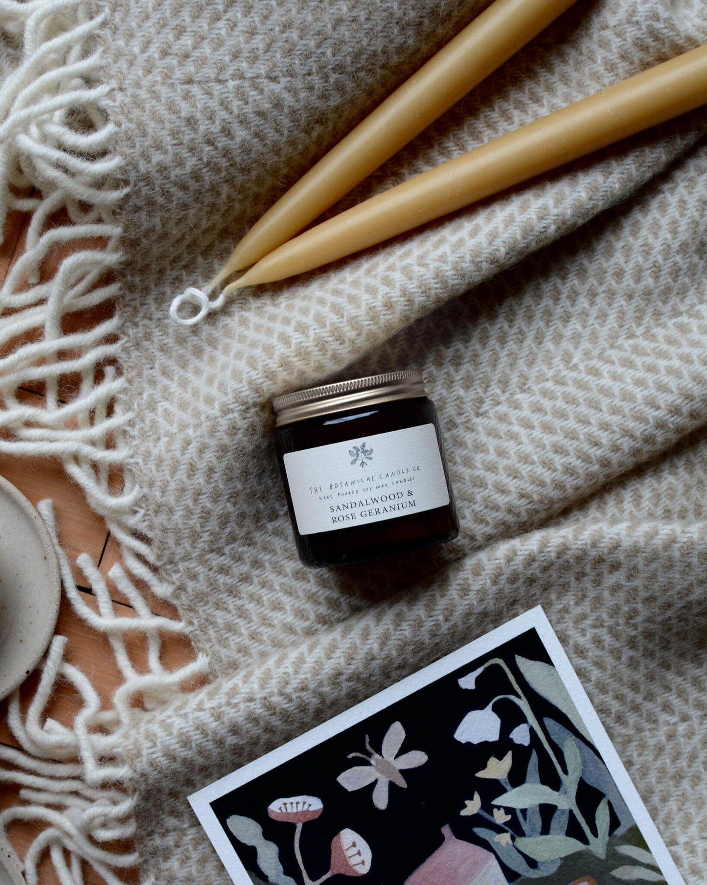 Sandalwood and Rose Geranium scented candle by The Botanical candle co. on a woollen blanket next to beeswax candles.