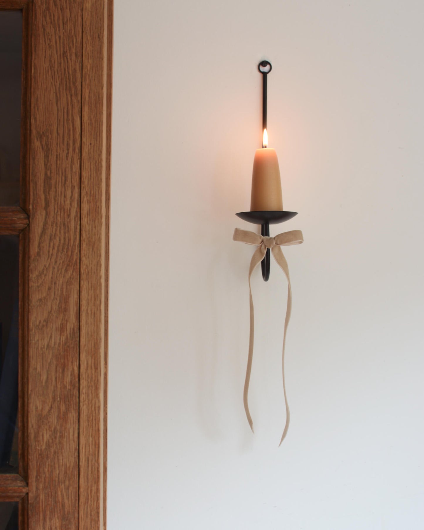 A single black metal wall sconce candle holder with a lit beeswax stubby candle and a velvet bow beneath.
