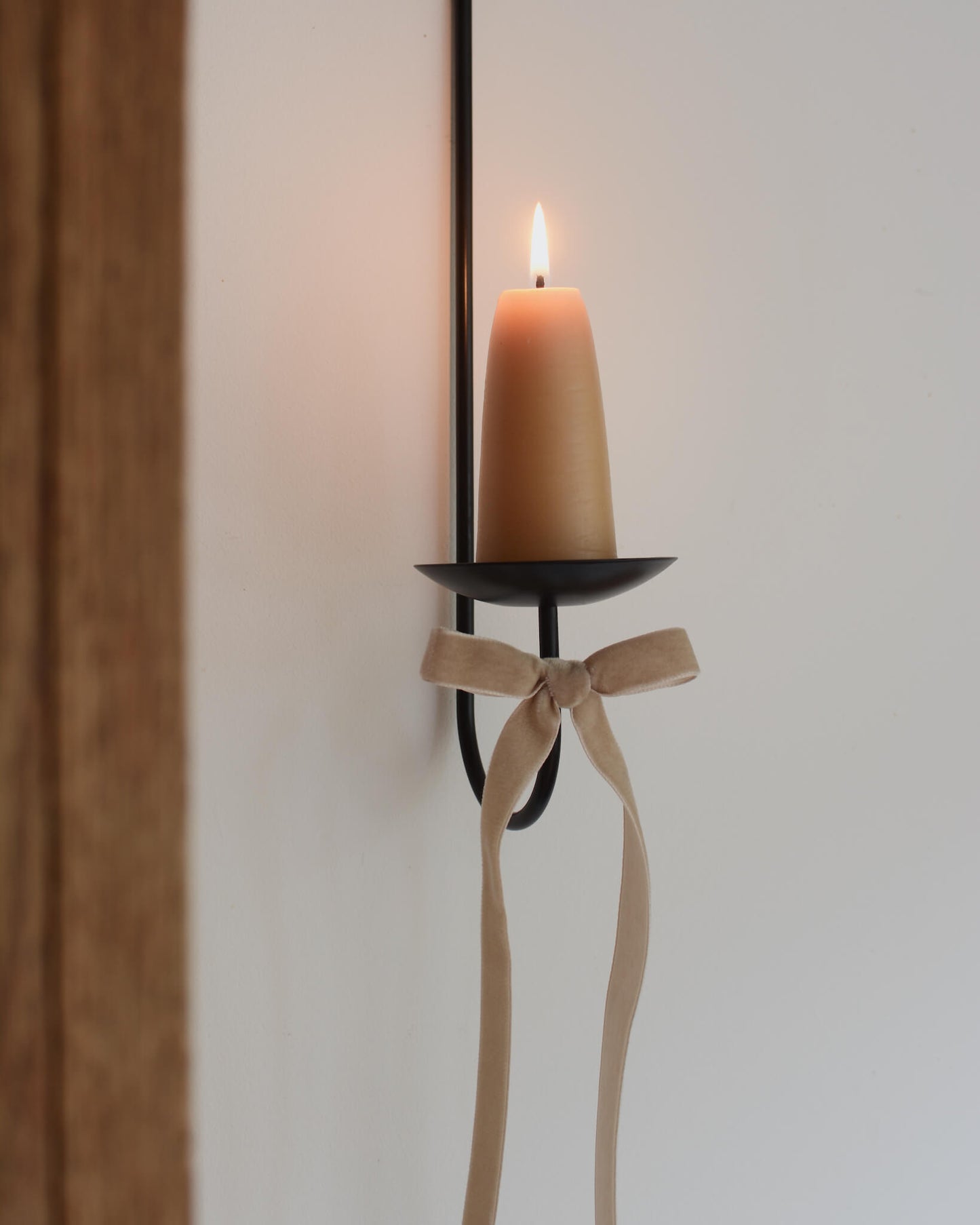 A single black metal wall sconce candle holder with a lit beeswax stubby candle and a velvet bow beneath.