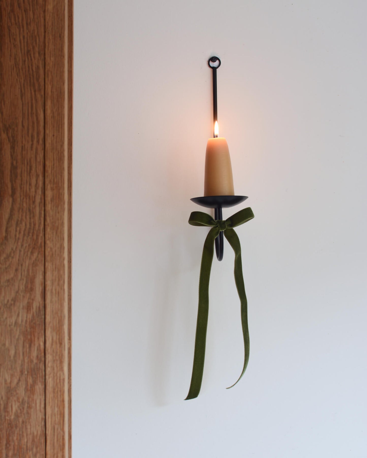 A single black metal wall sconce candle holder with a lit beeswax stubby candle and a velvet bow beneath.