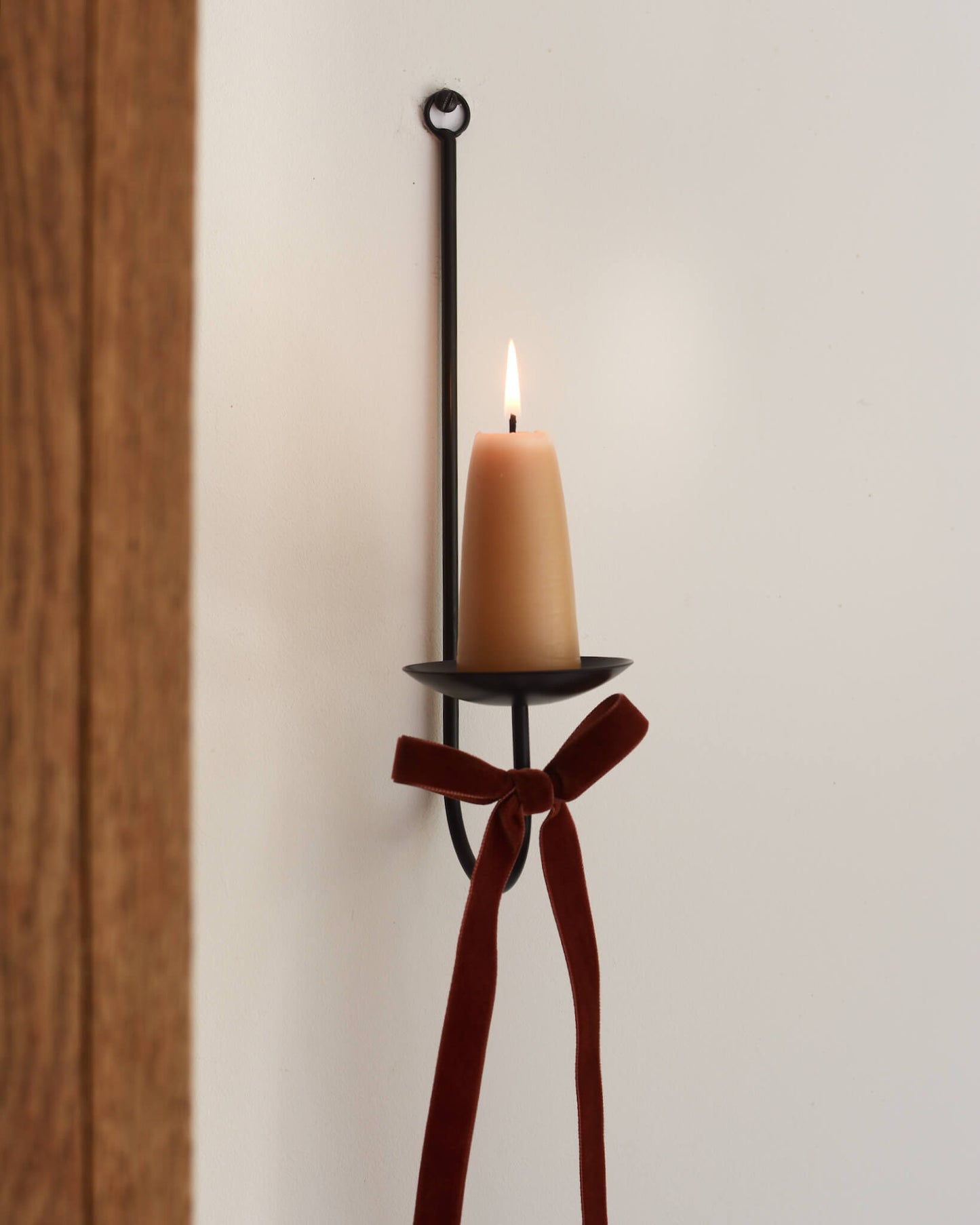 A single black metal wall sconce candle holder with a lit beeswax stubby candle and a velvet bow beneath.