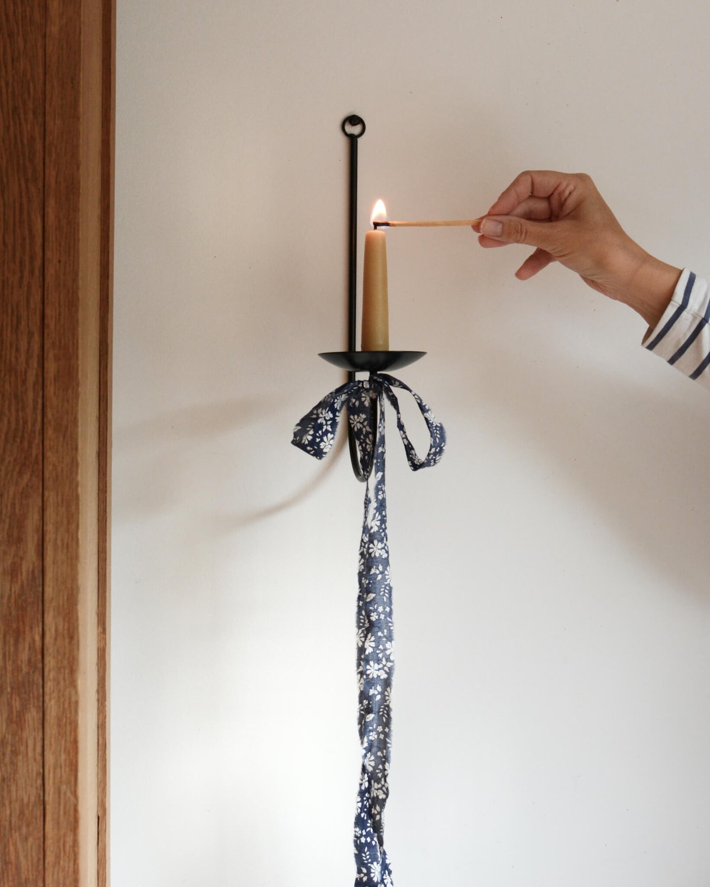 A single black metal wall sconce candle holder with a lit beeswax stubby candle and a floral bow beneath.