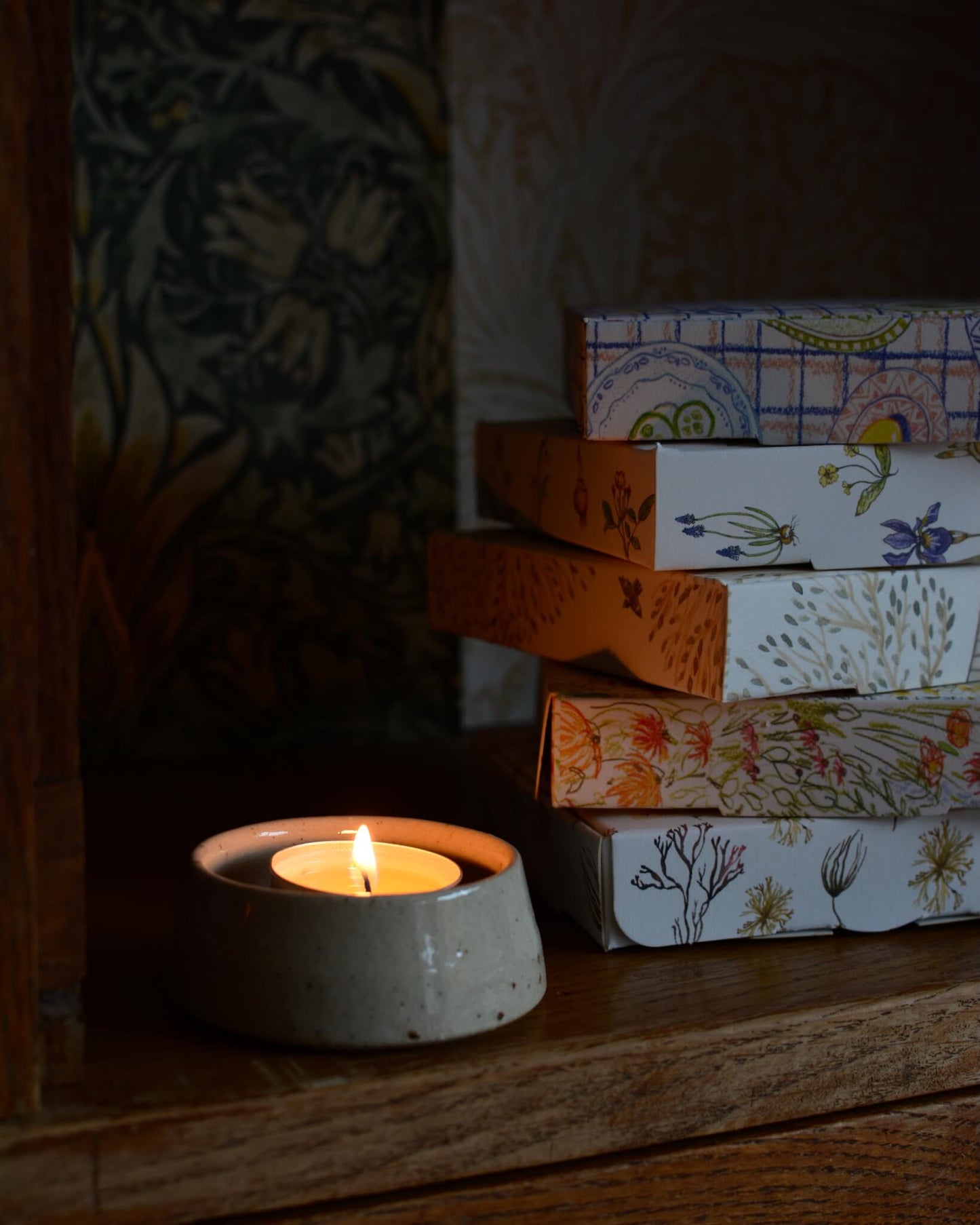 A lit tealight in a ceramic holder, next to a stack of illustrated tealight boxes