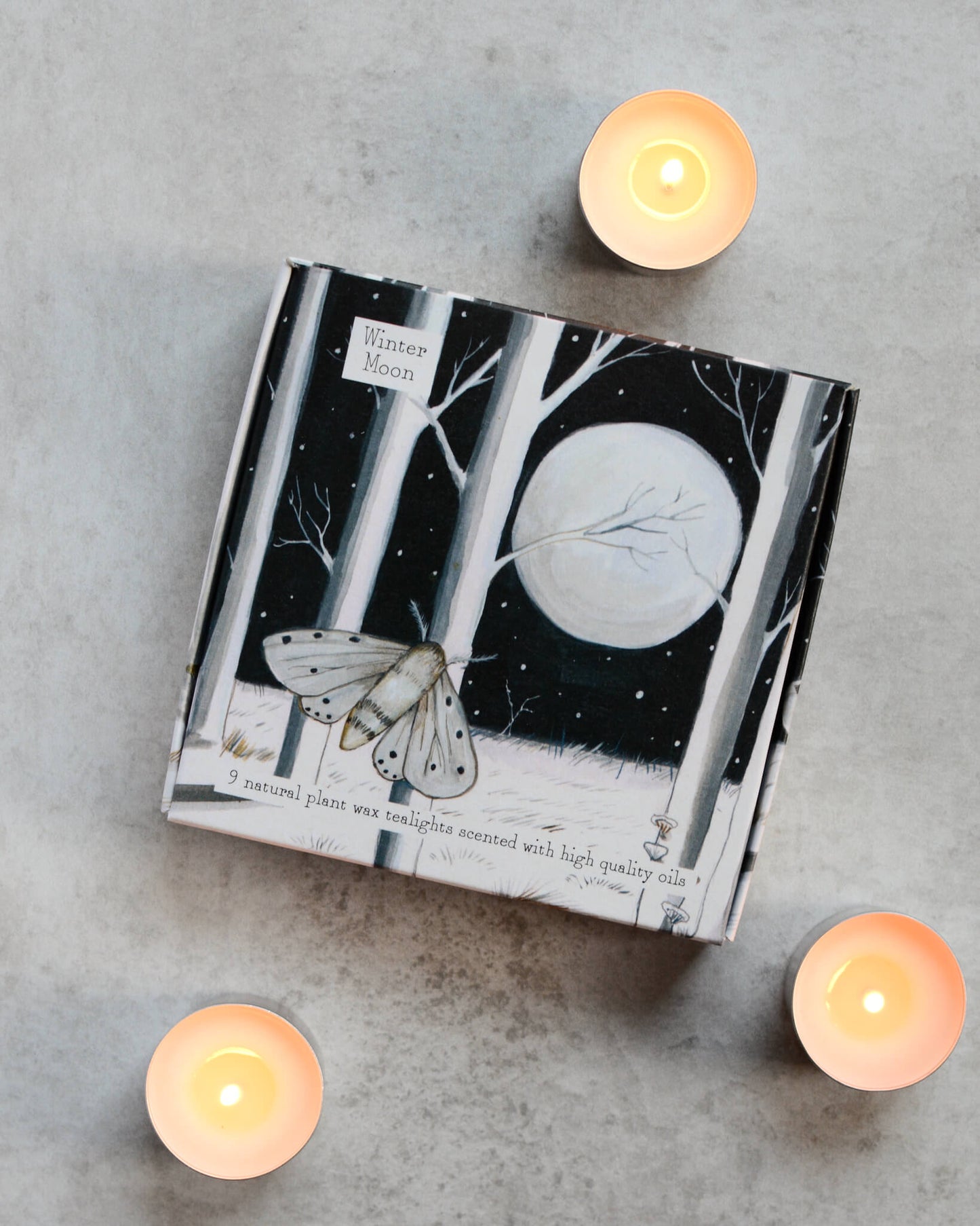 A set of lit tealights alongside a box with a monochrome forest and moon illustration.