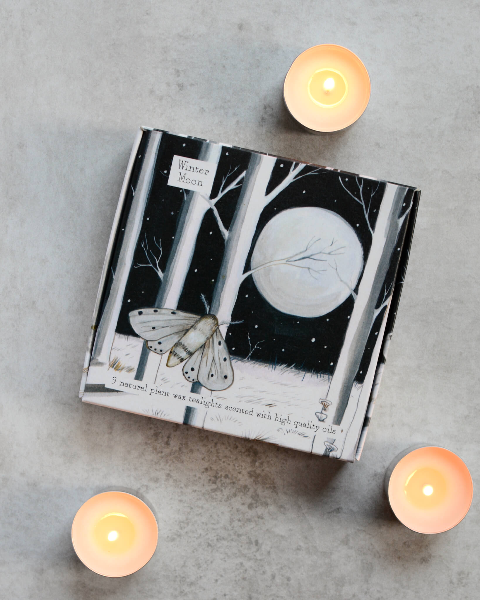 A set of lit tealights alongside a box with a monochrome forest and moon illustration.