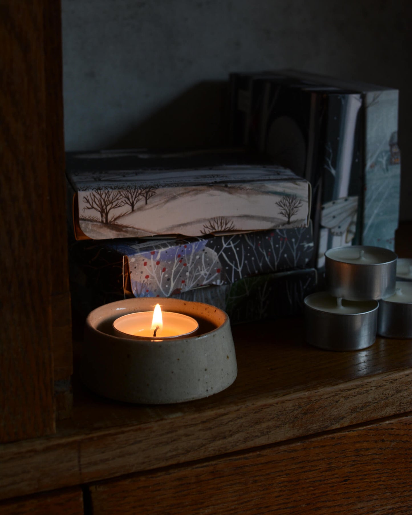 A lit tealight in a ceramic candle holder, n front of a stack of illustrated tealights boxes
