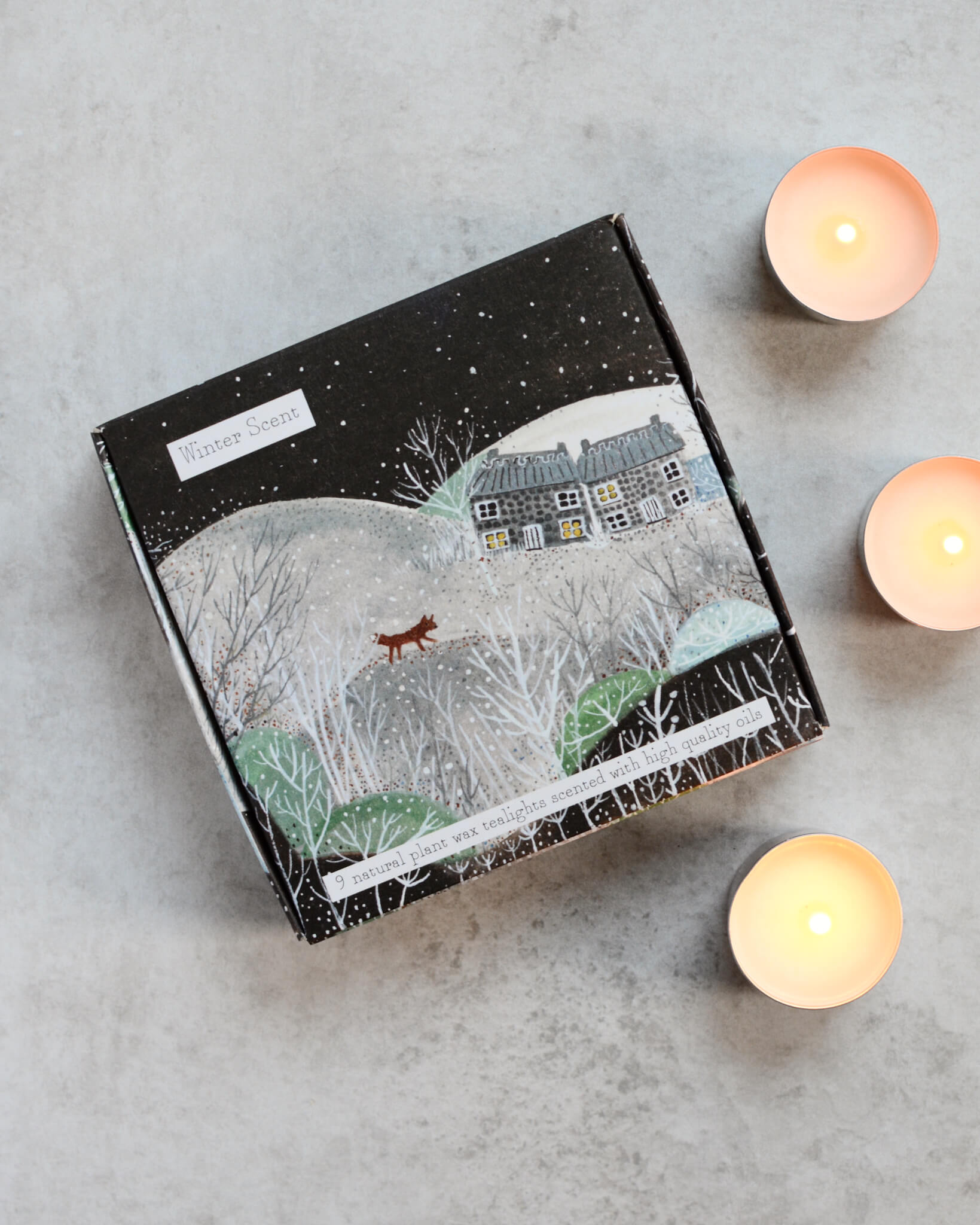 A box of tealights with a winter-themed design, including images of a fox and cottages, alongside lit tealights outside the box.
