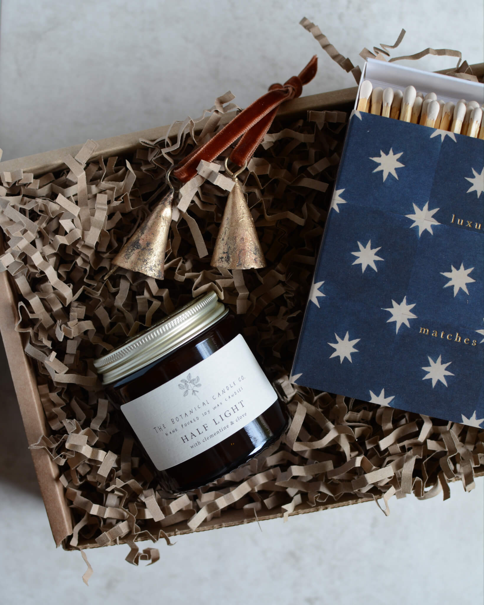A Christmas gift box containing a glass jar festive candle, a box of matches with a star design and a pair of brass bell ornaments.