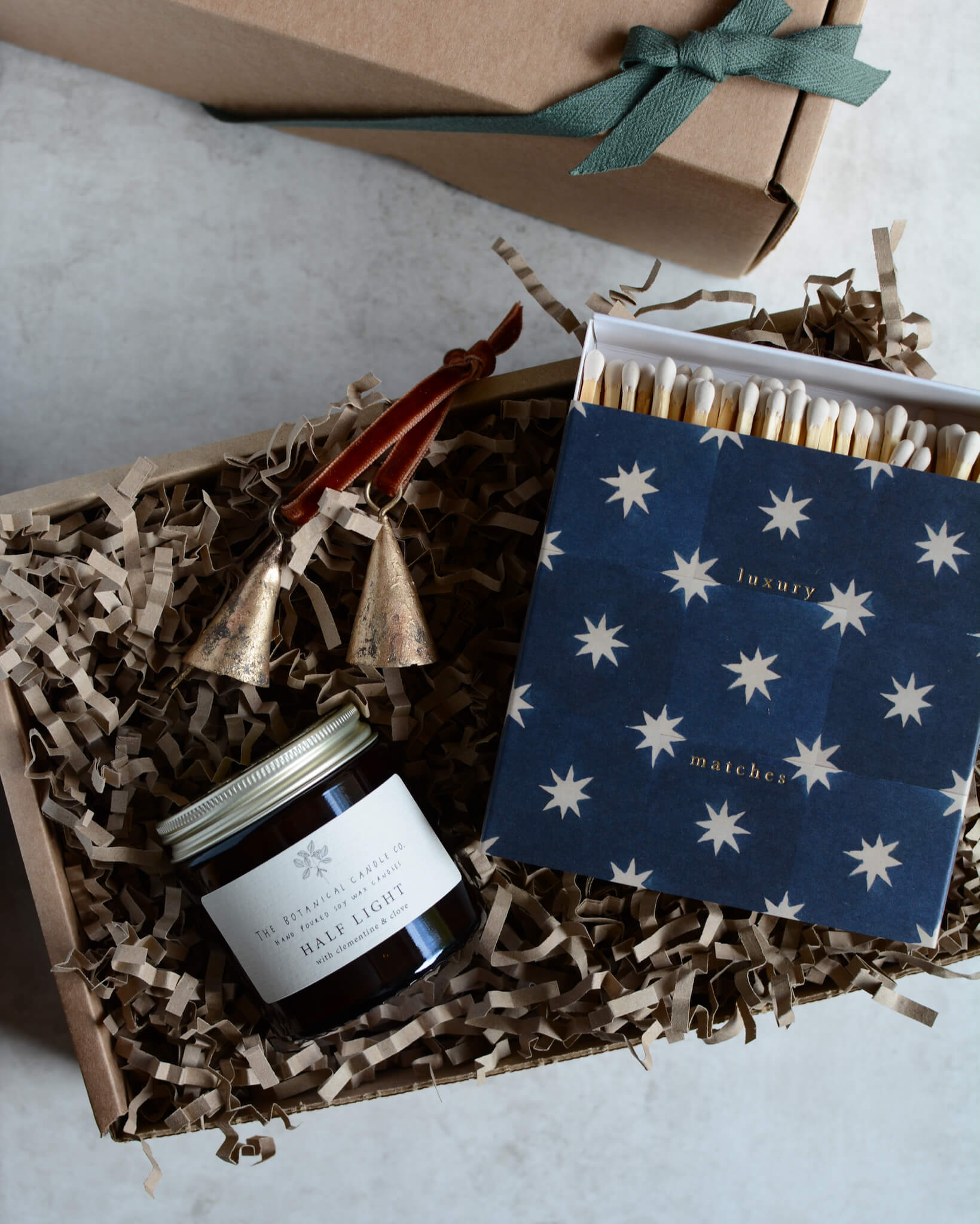 A Christmas gift box containing a glass jar festive candle, a box of matches with a star design and a pair of brass bell ornaments.
