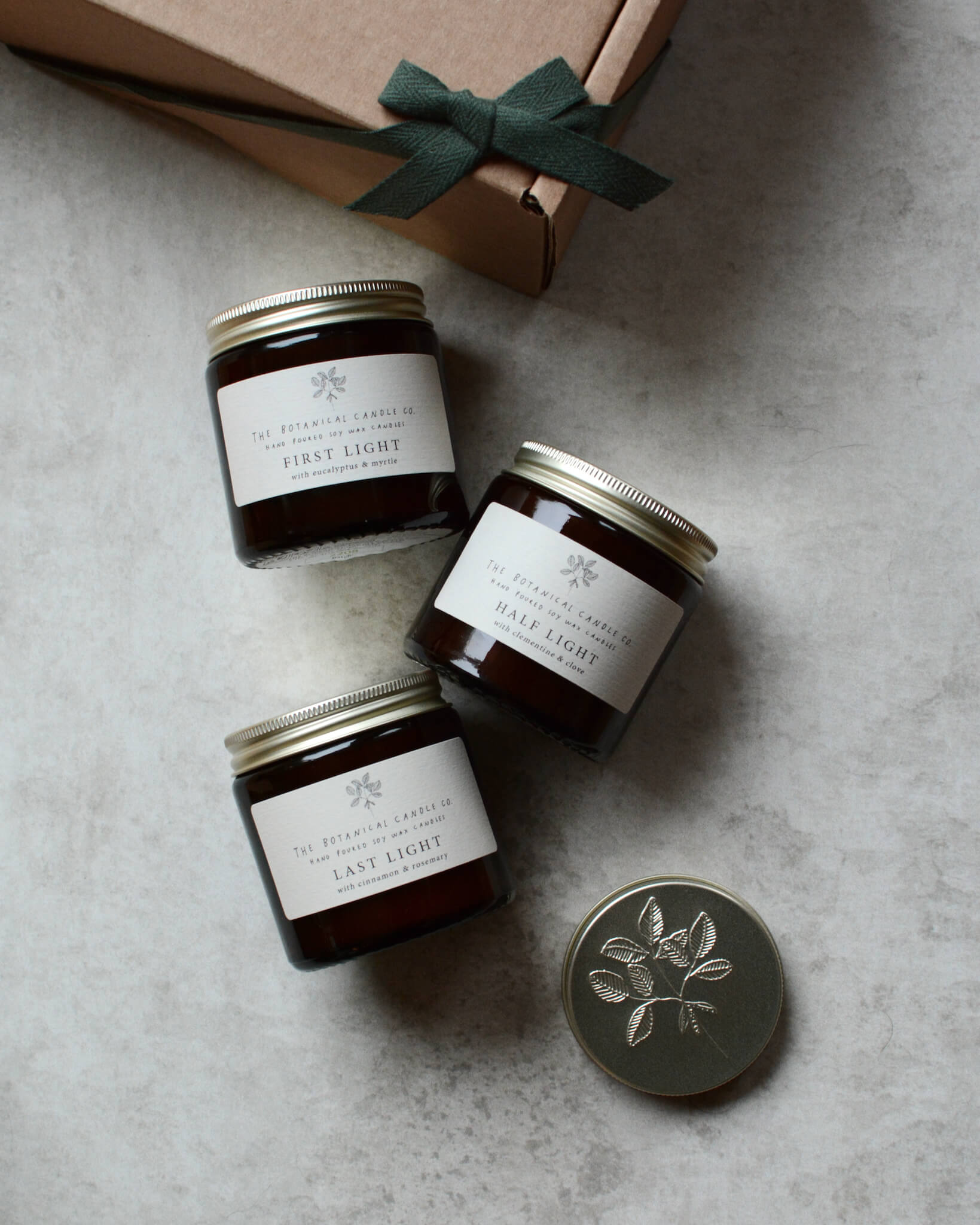 A trio of festive candles in glass jars, shown next to a gift box.