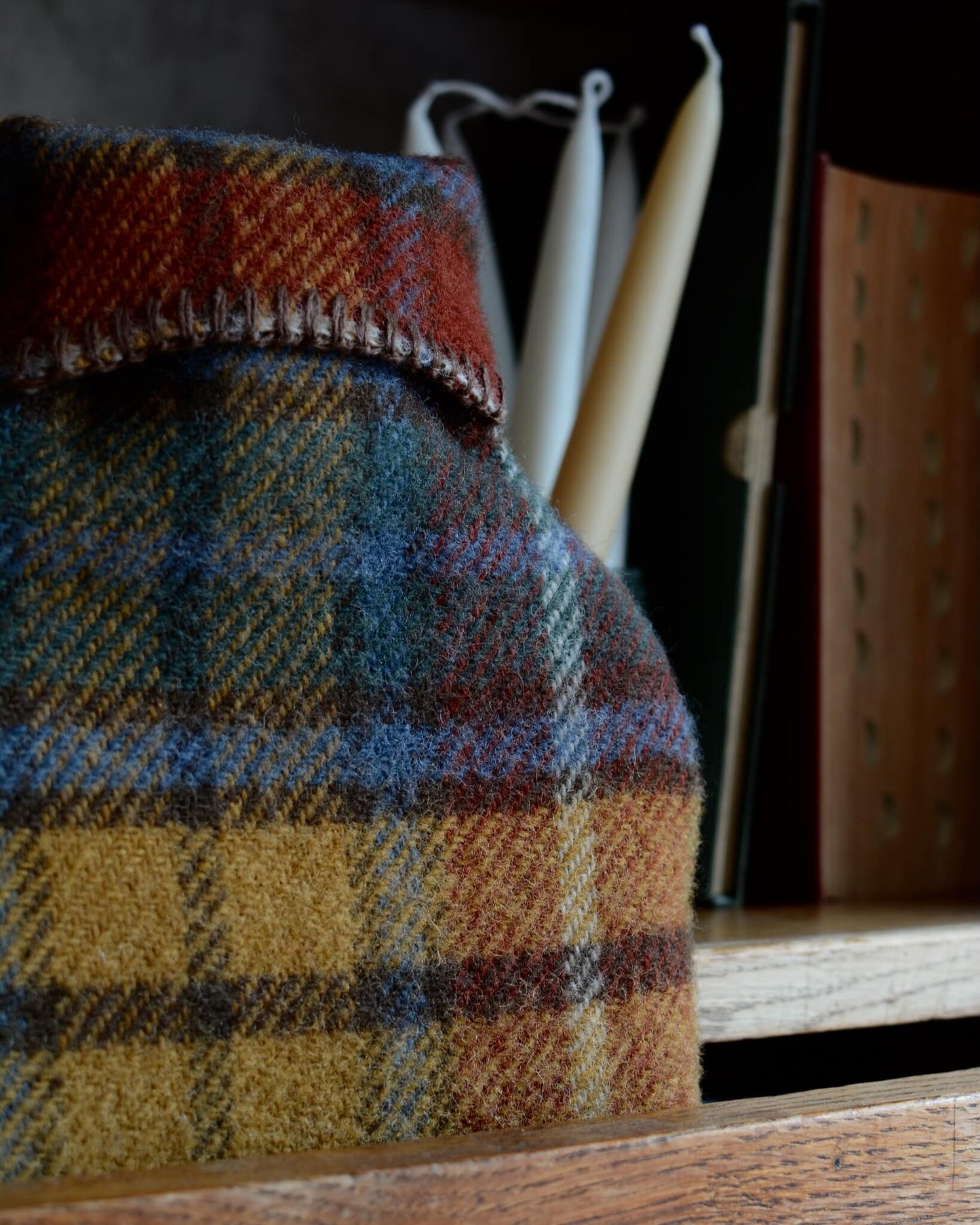Fireside tartan wool hot water bottle cover by Tweedmill Textiles, in ochre, rust and teal plaid, shown on a wooden drawer unit.