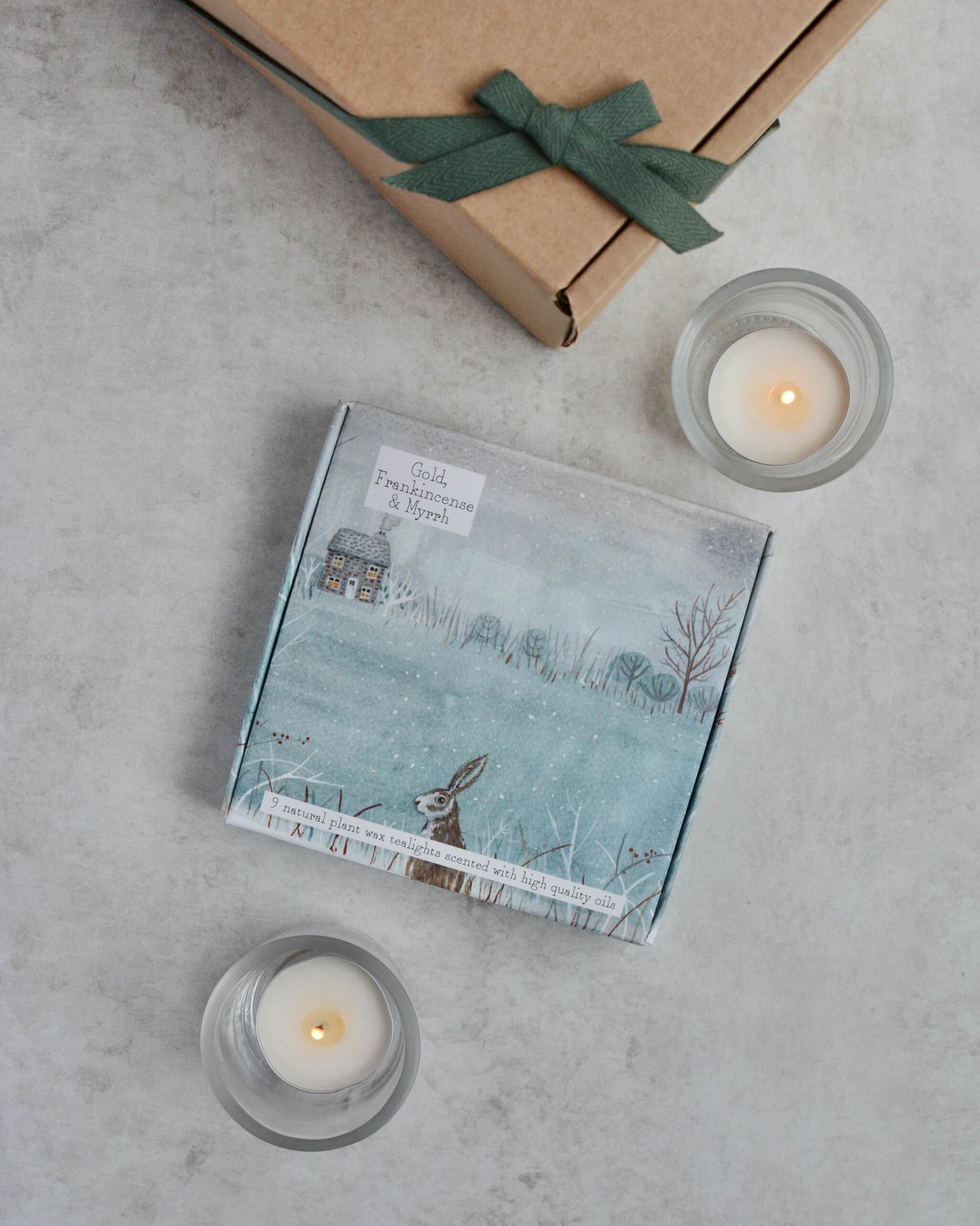Box of tealights with a winter landscape illustration, two lit tealight candles, and a brown box with a green ribbon on a grey surface.