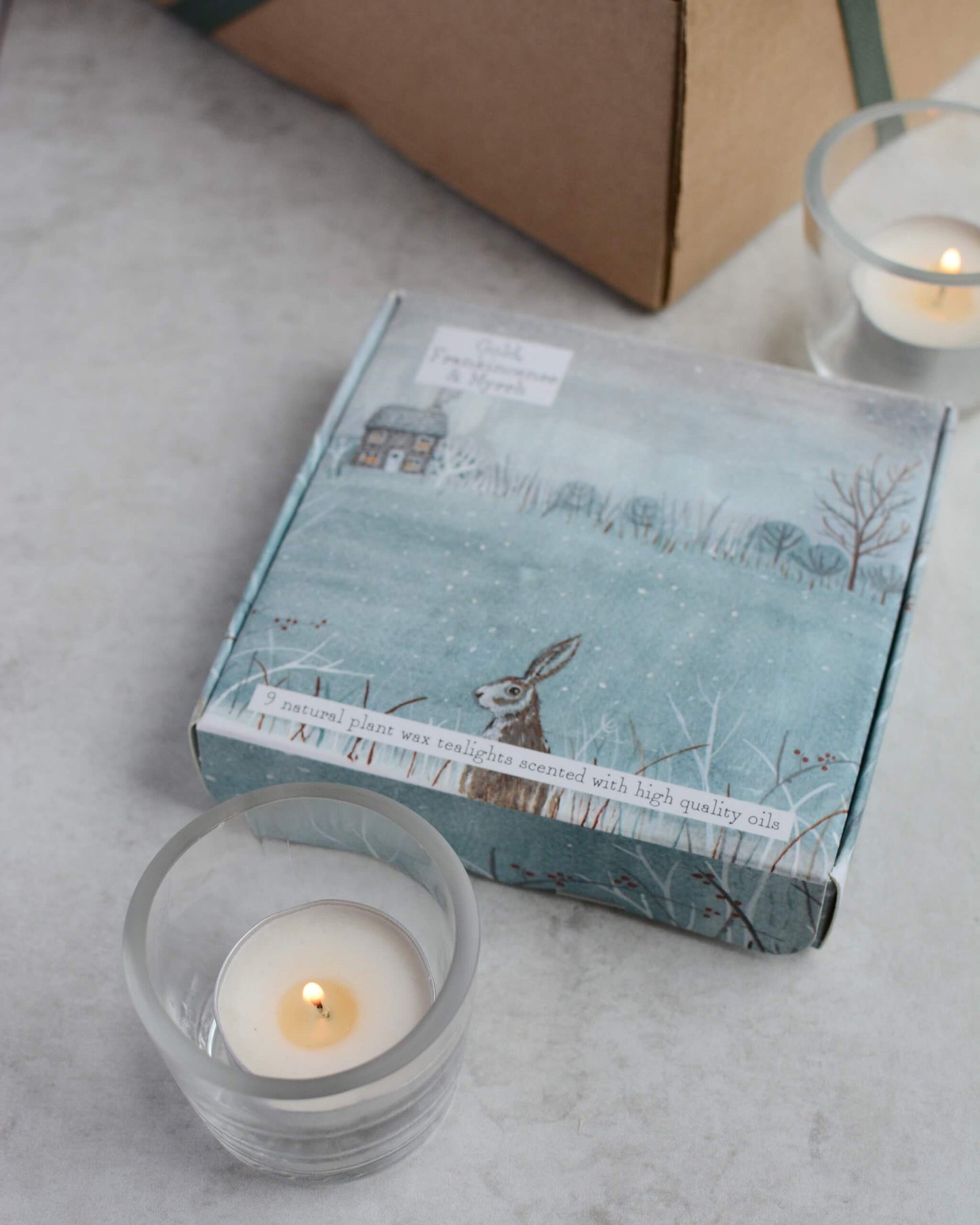 Box of tealights with winter scene design and lit candles on a light surface.