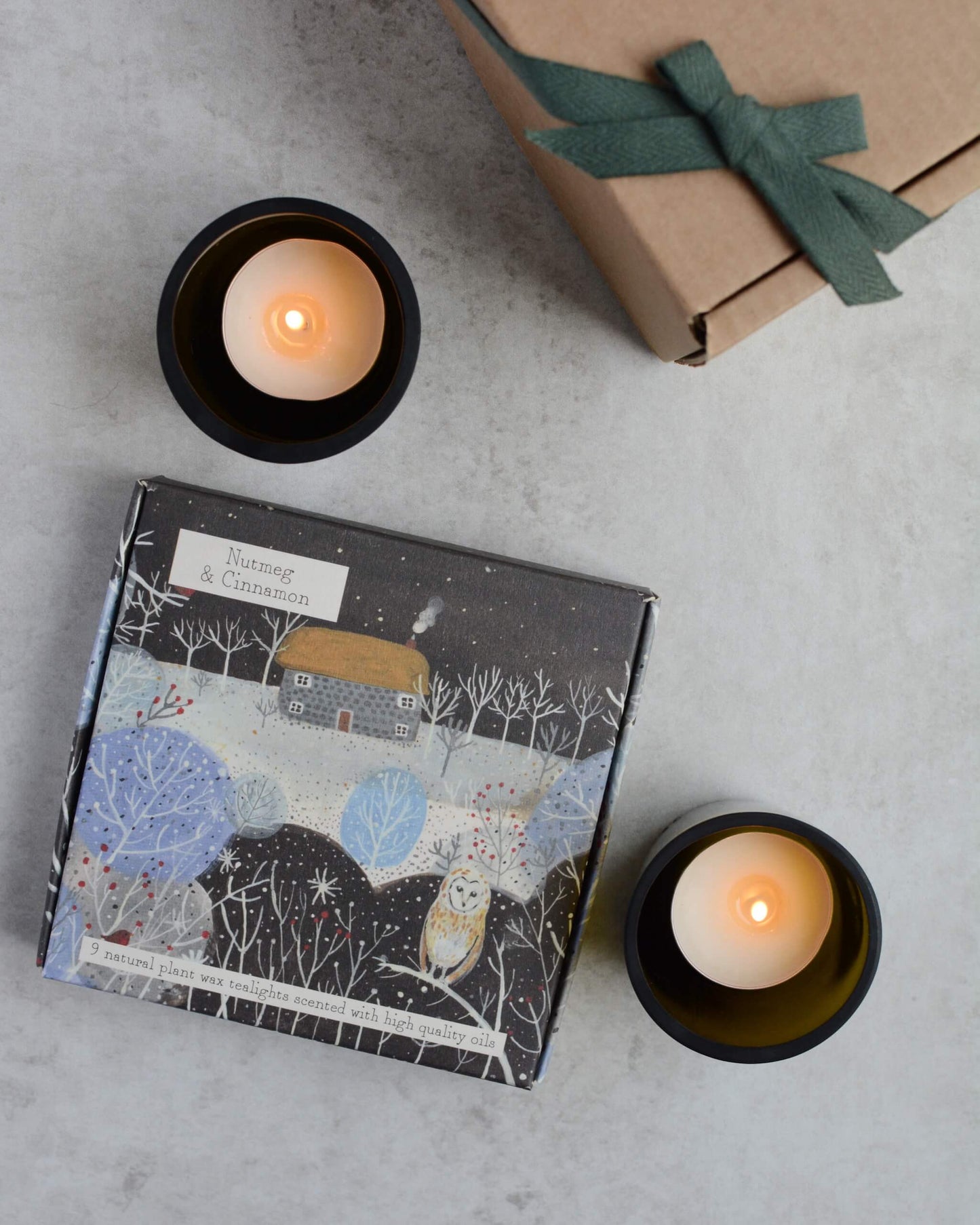 Two lit candles and a box of illustrated tealights on a grey surface.