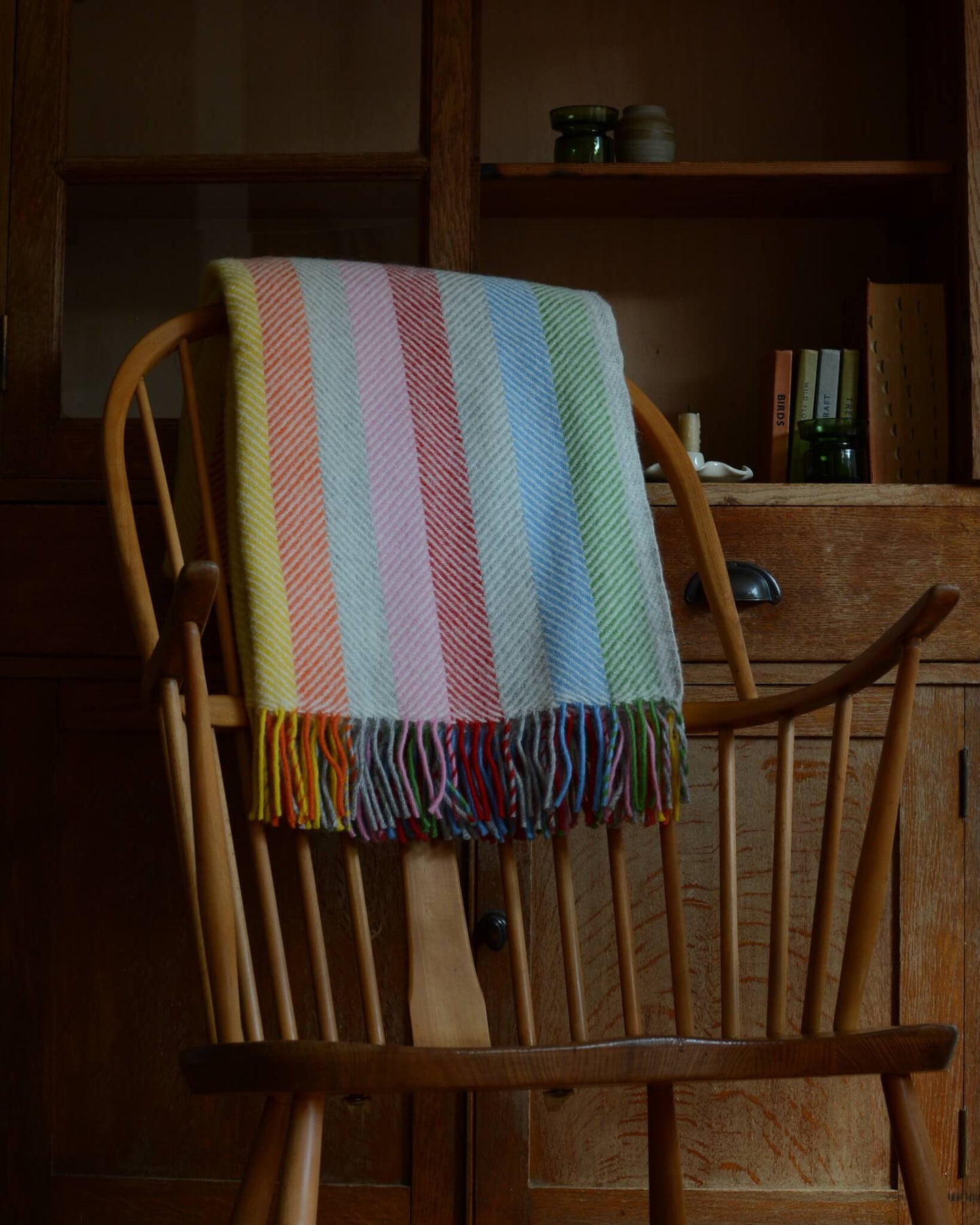 Colourful rainbow striped wool blanket with fringe, shown on a wooden rocking chair.