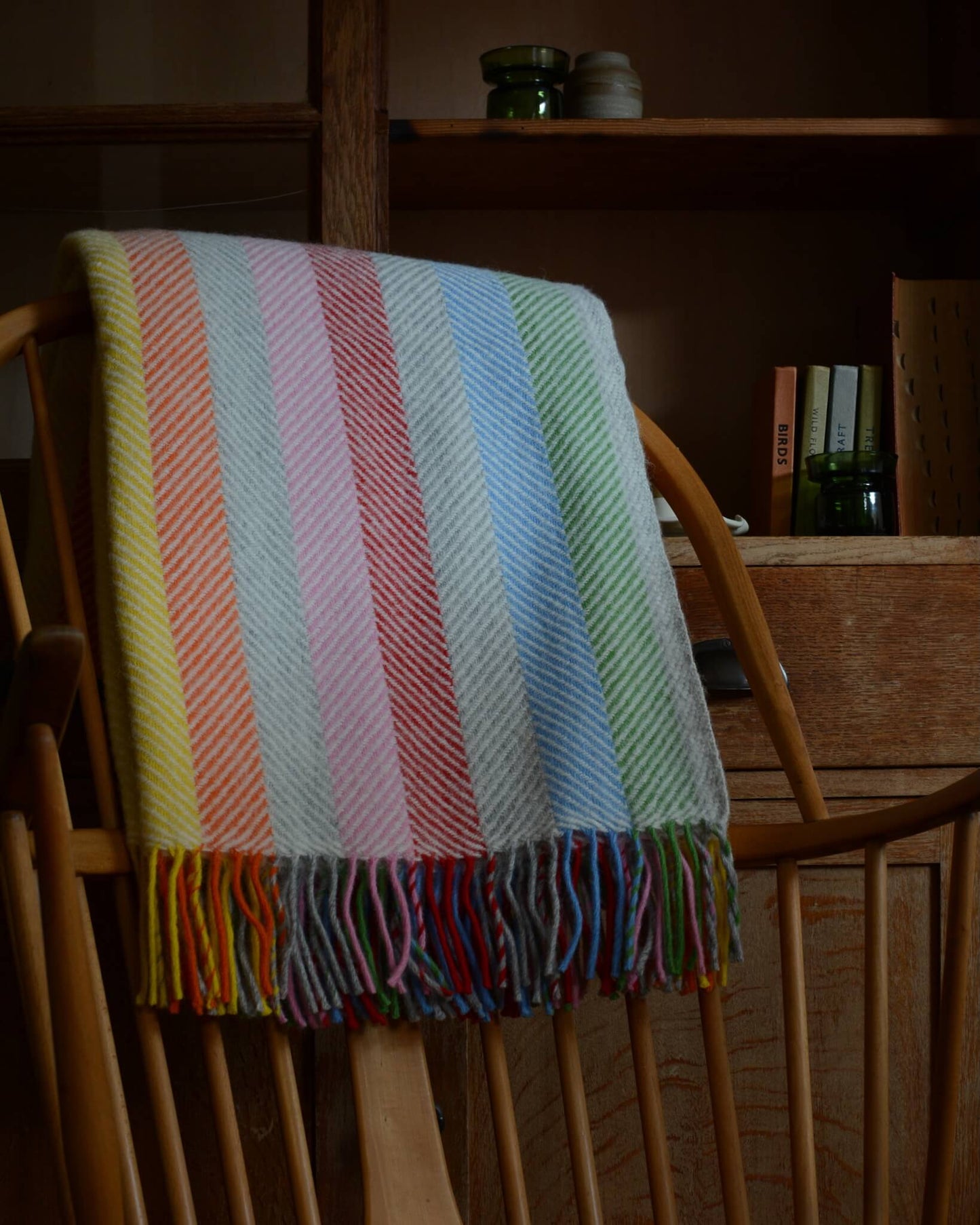 Colourful rainbow striped wool blanket with fringe, shown on a wooden rocking chair.