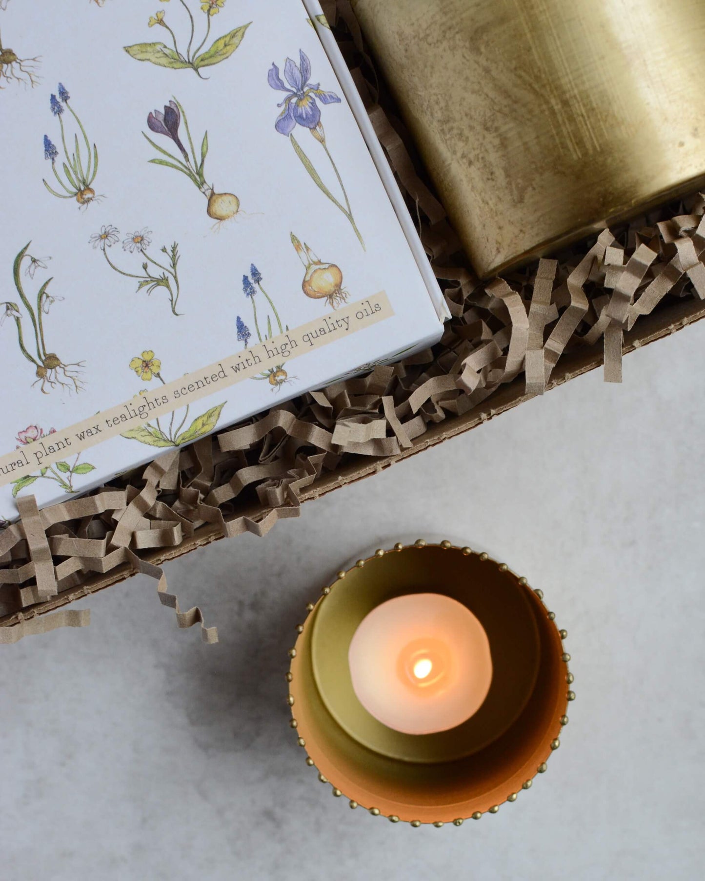 Tealight candle in a decorative brass holder with a box featuring botanical illustrations on a light surface.