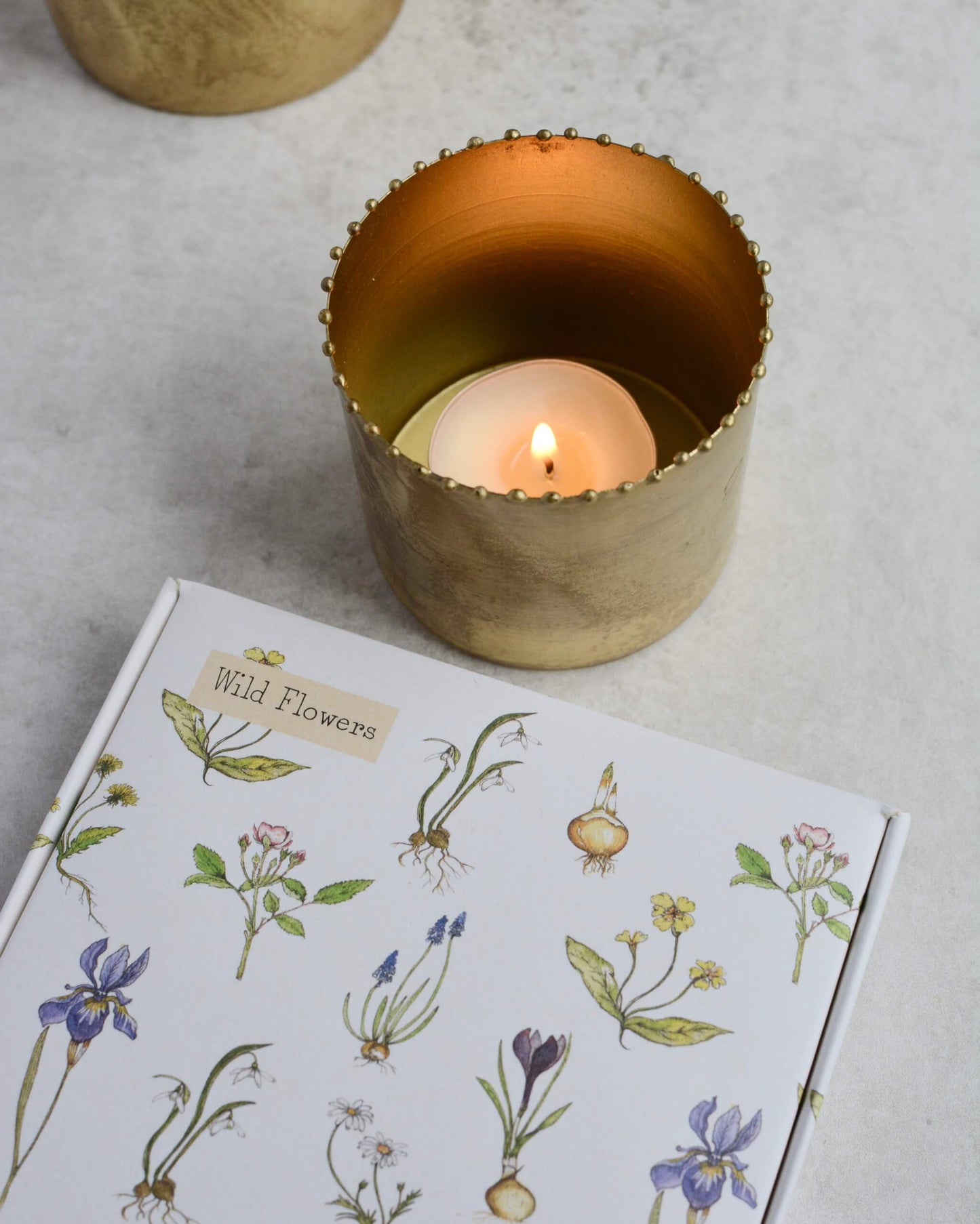 A lit tealight within a brass holder, shown next to an illustrated box of scented tealights.