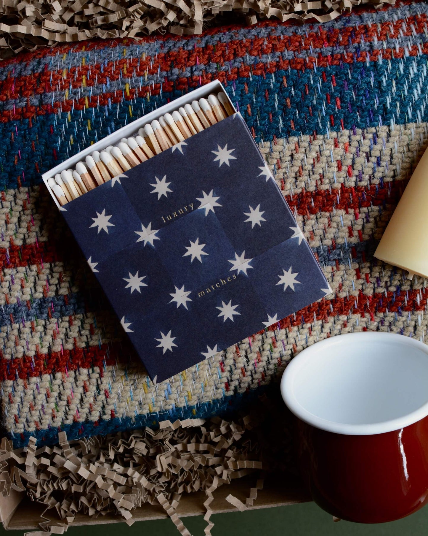 A gift box containing a wool blanket, box of matches, red enamel mug and pair of stubby beeswax candles.