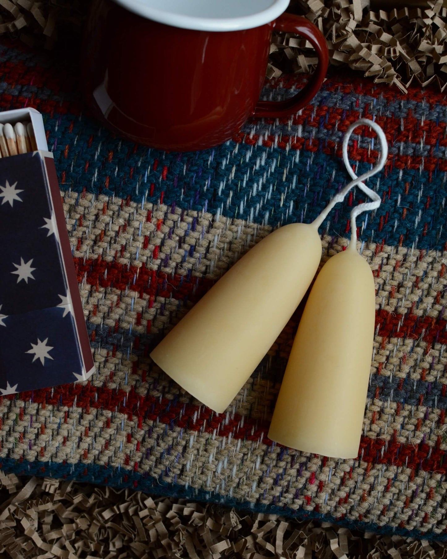 A gift box containing a wool blanket, box of matches, red enamel mug and pair of stubby beeswax candles.