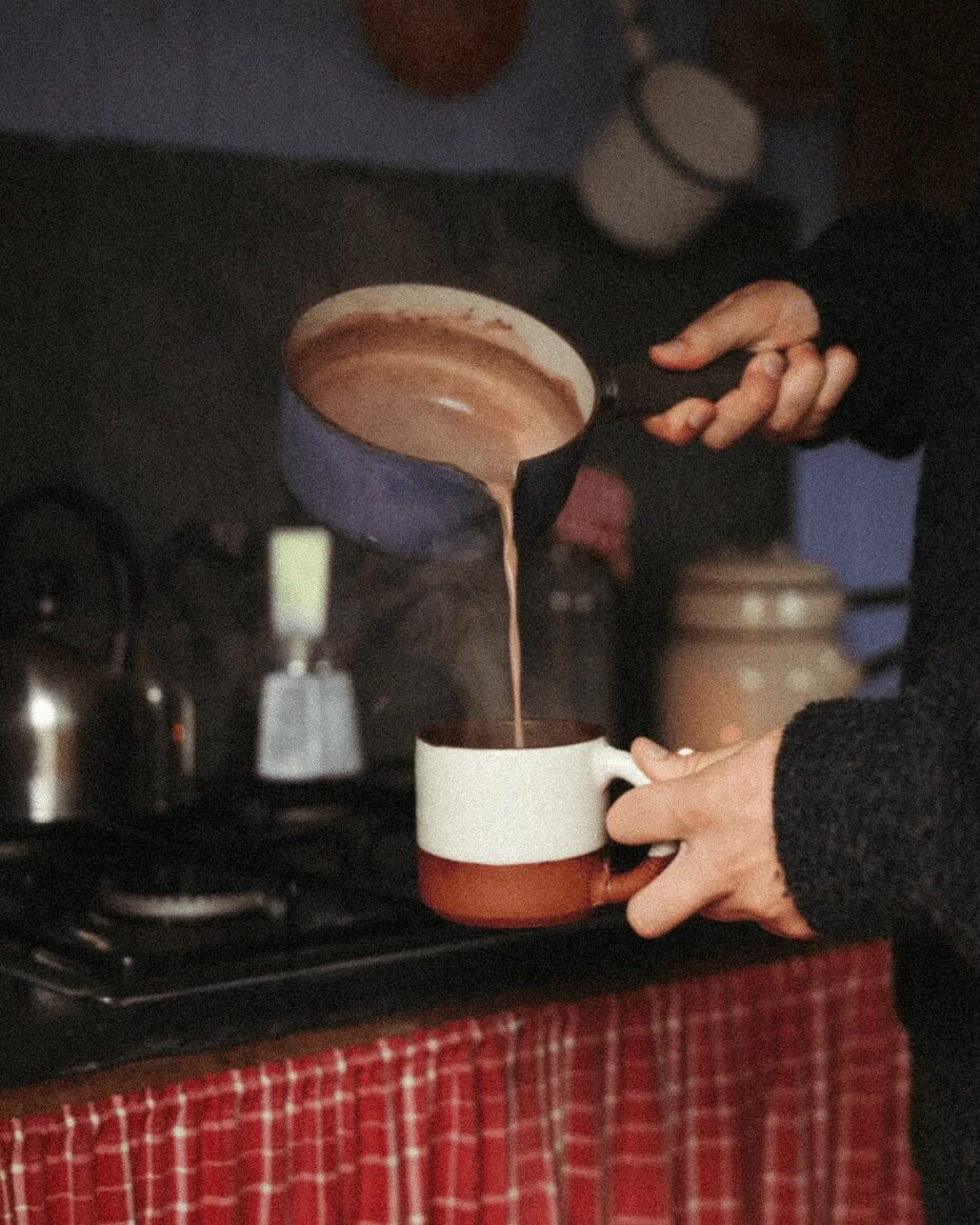 Person pouring hot chocolate from a saucepan into a mug on a stove.