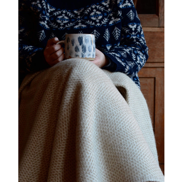 Tweedmill oatmeal beehive wool throw shown draped over the lap of a woman holding a coffee cup.