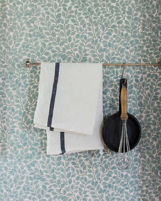 A wall with wallpaper and a brass rail holding a pan, whisk and kitchen linens.