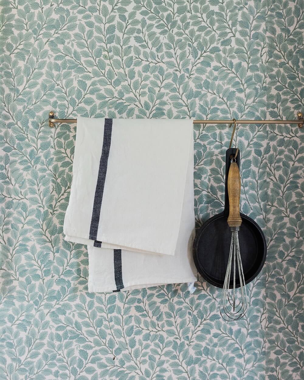 A wall with wallpaper and a brass rail holding a pan, whisk and kitchen linens.