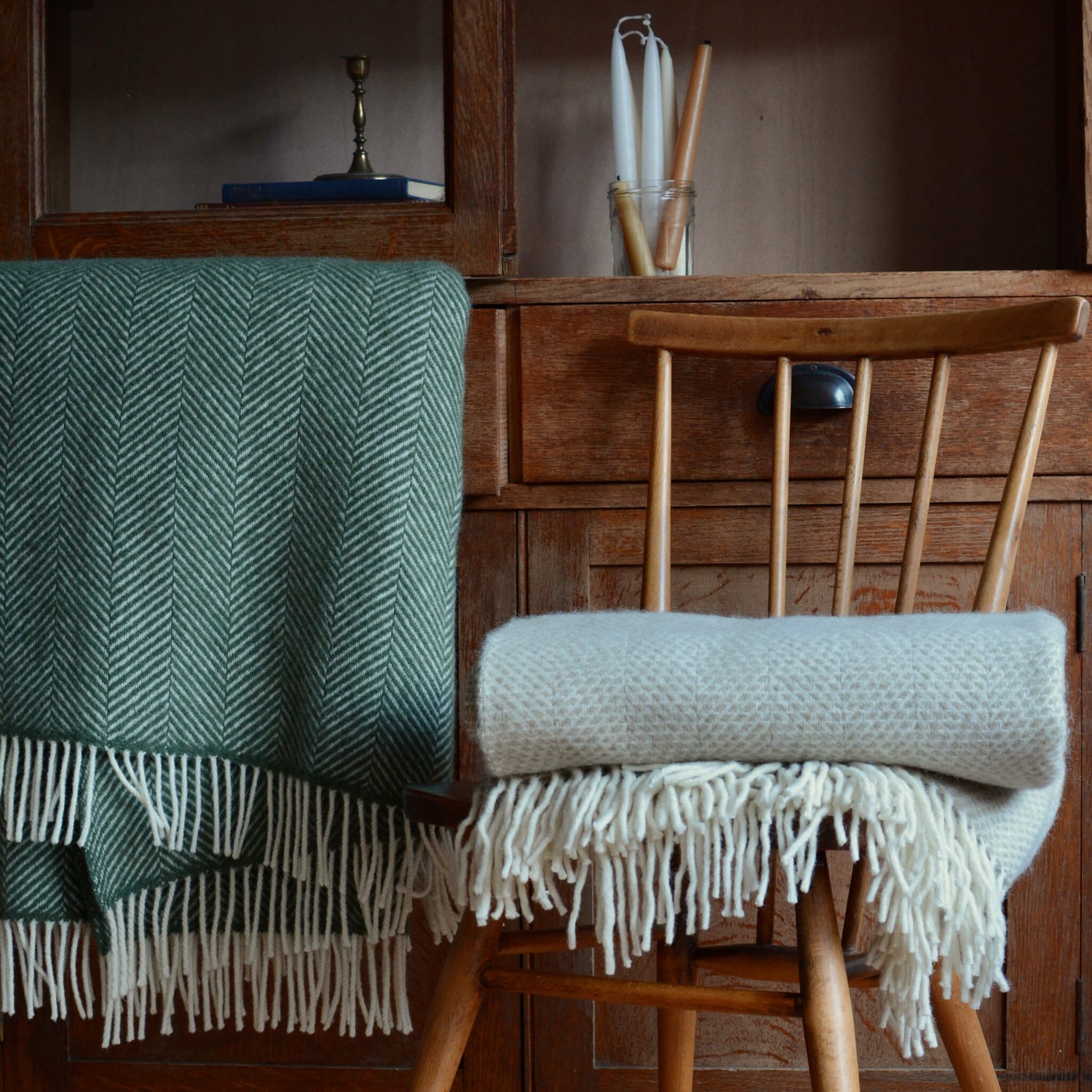 A green and oatmeal wool blanket with fringe. Infront of a wooden cabinet and resting on a wooden chair.