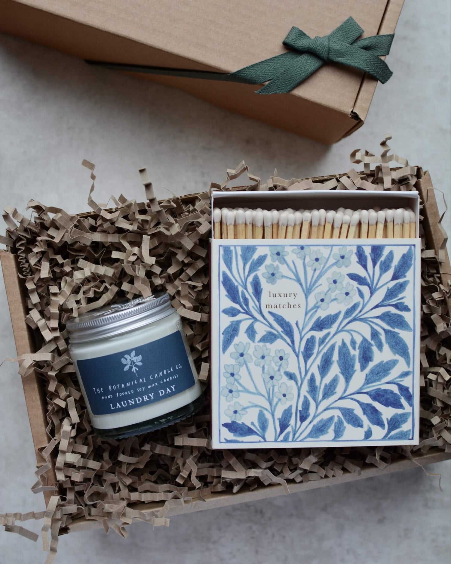 A gift box containing a glass jar candle and a blue floral box of matches.
