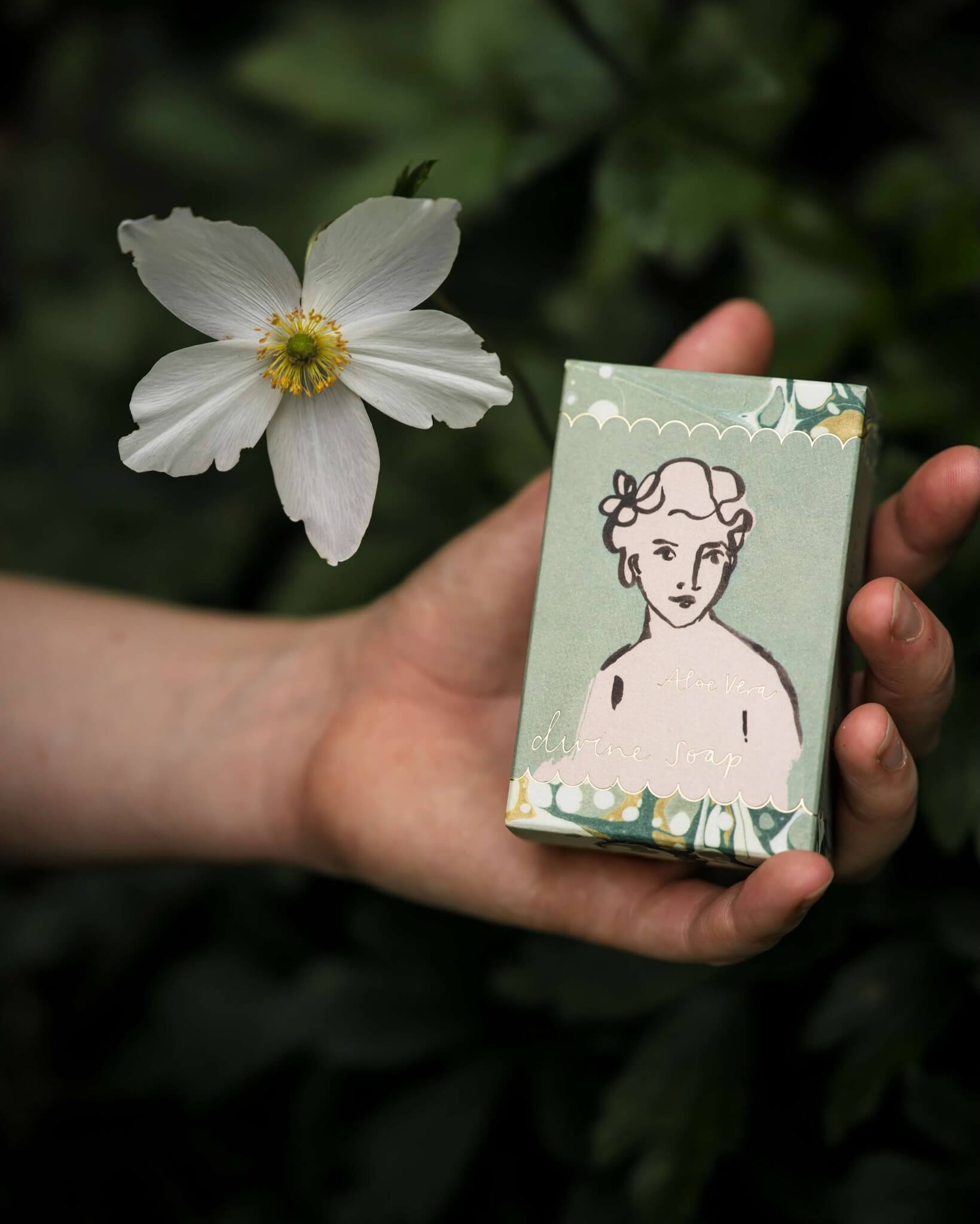Hand holding a green box of soap with a printed design of a woman's face, next to a white flower against a green background.